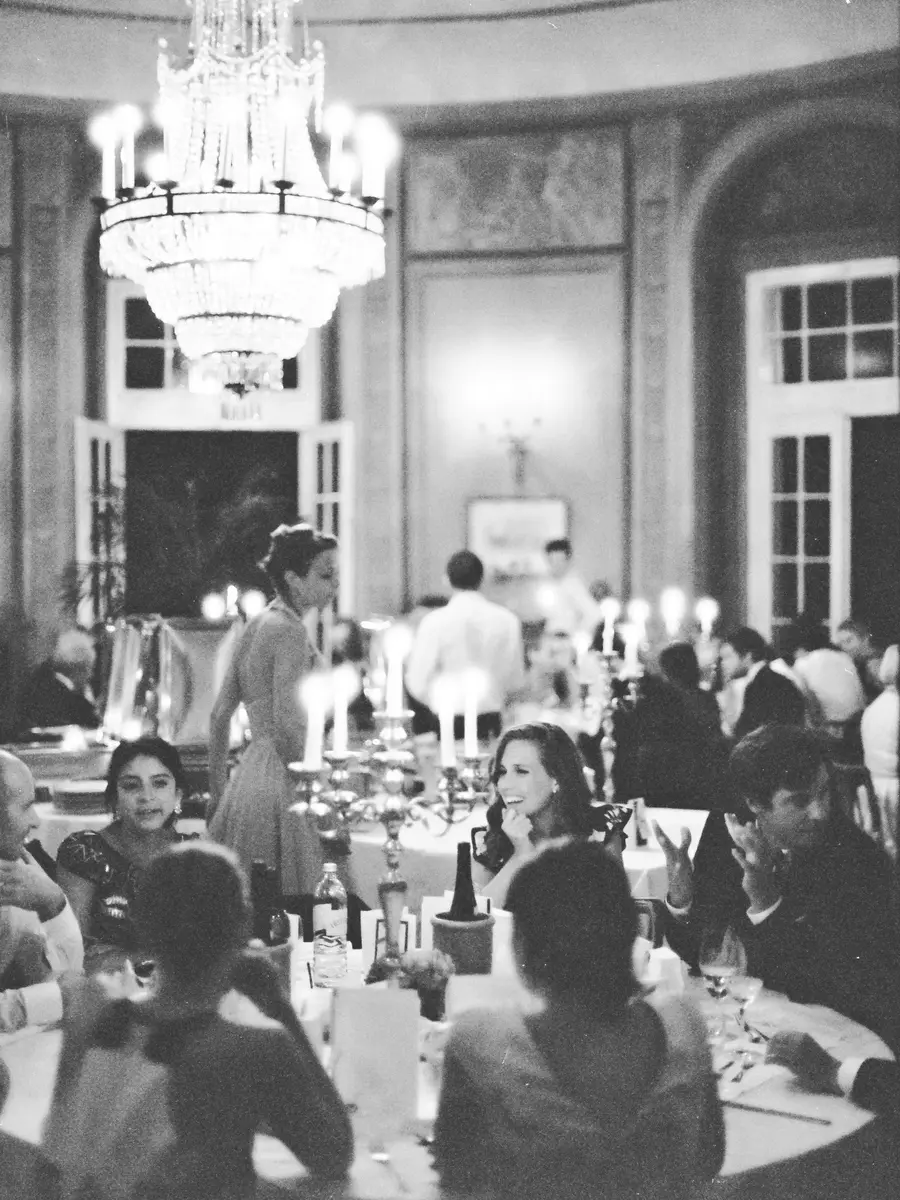 Atmospheric wedding formal dinner shot on Ilford Delta 3200. Several tables are set with elaborate candelabras and glassware. In the foreground, guests are seated at a table, with a woman on the right laughing. Behind them, other guests and staff are visible, along with a grand, ornate chandelier hanging from the ceiling.