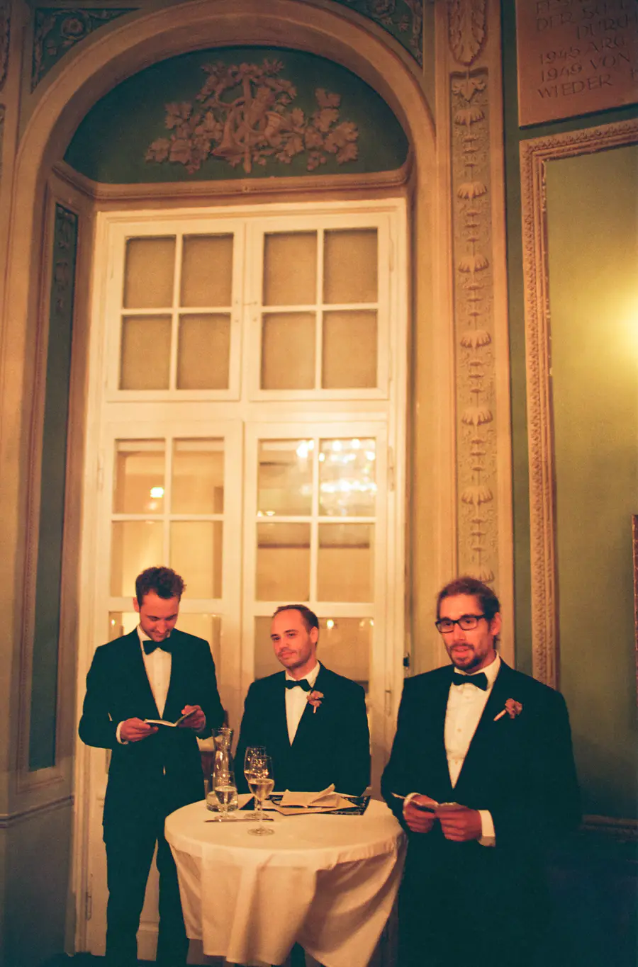 Three men in tuxedos stand before a large window. The man on the left holds a program and looks down at it, the man in the middle looks towards the right with a slight smile, and the man on the right wears glasses and holds a program in his hands.