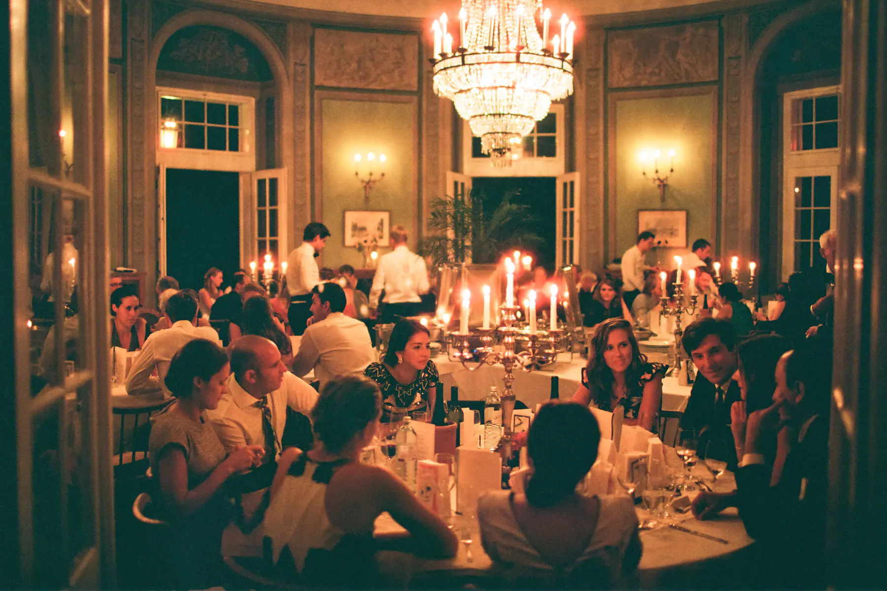 Warm evening dinner captured on Kodak Portra 800 film – authentic natural light photography. Lavishly decorated room, illuminated by a large, ornate chandelier and numerous candles. Guests, dressed in elegant attire, are seated at long tables laden with food and drink, engaged in conversation and enjoying the festive atmosphere. The room features high ceilings, arched windows, and decorative wall moldings, contributing to the opulent ambiance of the event.