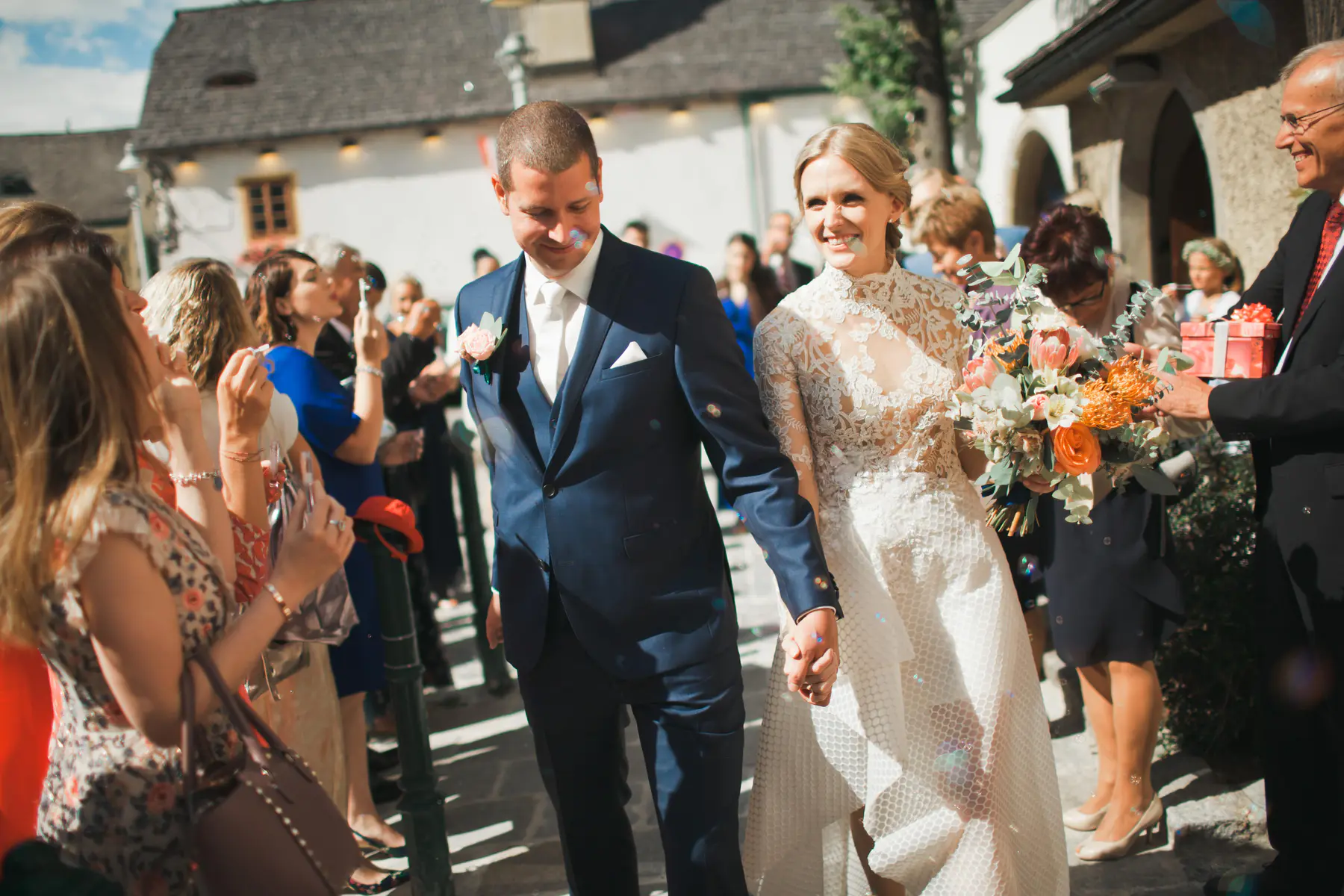 A smiling bride in a white lace dress, holding a colorful bouquet, walks hand-in-hand with her groom in a dark suit down a cobblestone path. Guests line the path, blowing bubbles