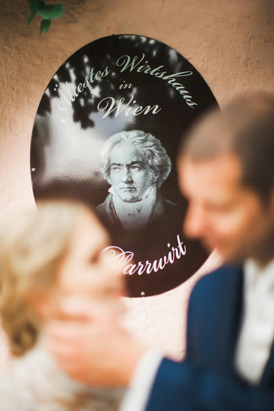 Künstlerische Hochzeitsfotografie Wien: Historisches Beethoven-Schild beim Pfarrwirt im Fokus, während das Brautpaar im Vordergrund bewusst unscharf bleibt. Kreative Bildkomposition und exklusives Storytelling im 19. Bezirk (Döbling). Dokumentarischer Stil von Marek & Klára