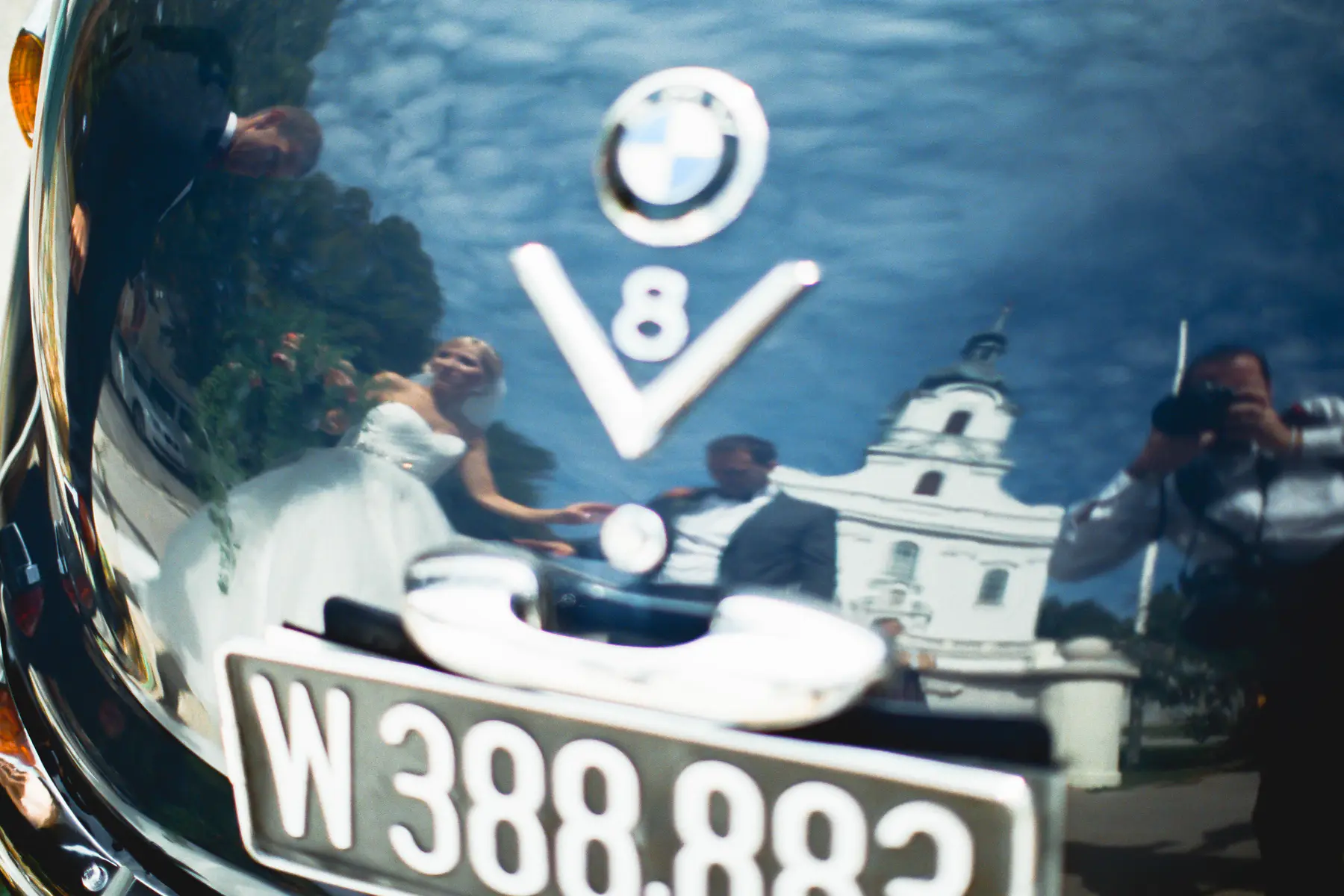 A wedding party is reflected in the shiny hood of a car. The bride and groom are in the foreground. The bride is wearing a white wedding dress, and the groom is wearing a dark suit. A BMW logo and "V8" are visible on the car's hood. A white church is in the background.