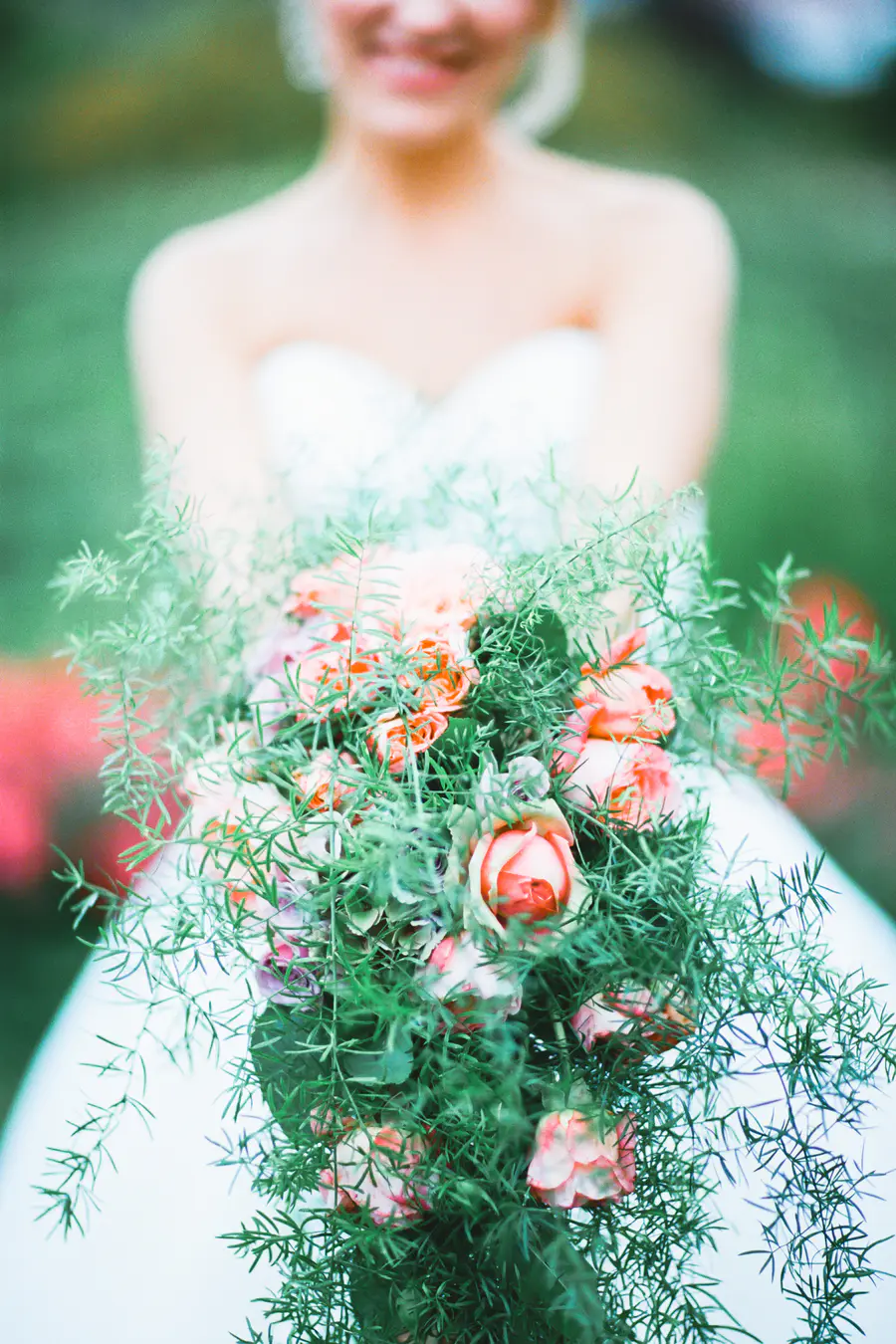 A smiling bride in a white strapless dress holds out a vibrant bouquet of peach and pink roses intertwined with abundant feathery green foliage. Her face is softly blurred in the background.Captured on Fuji Film PRO 400H