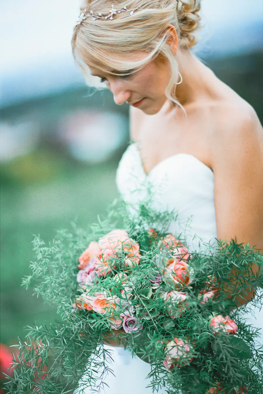 Captured on Fuji Film PRO 400H.A blonde bride in a white strapless gown and silver headpiece looks down at a large bouquet of peach, pink, and purple roses surrounded by abundant green foliage.