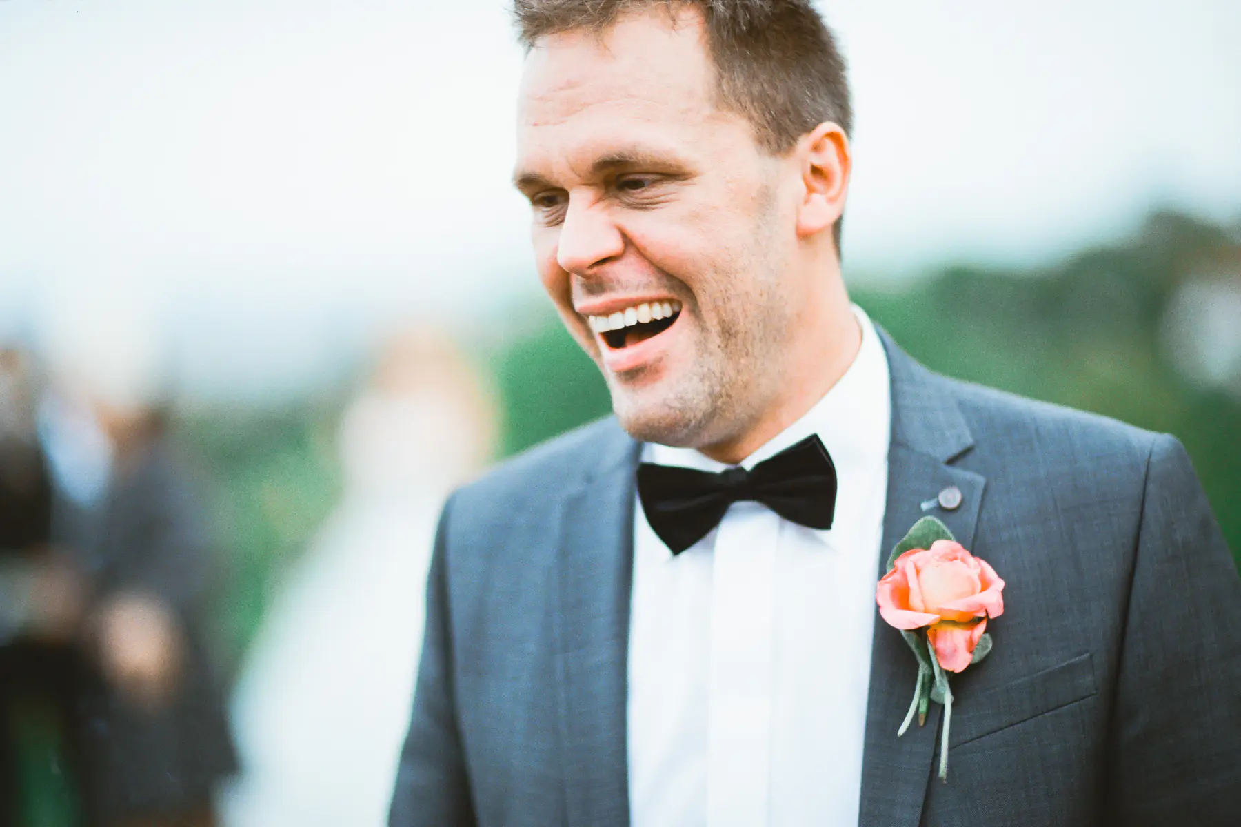 Captured on Fuji Film PRO 400H.A man in a grey suit, black bow tie, and peach rose boutonnière laughs heartily outdoors.