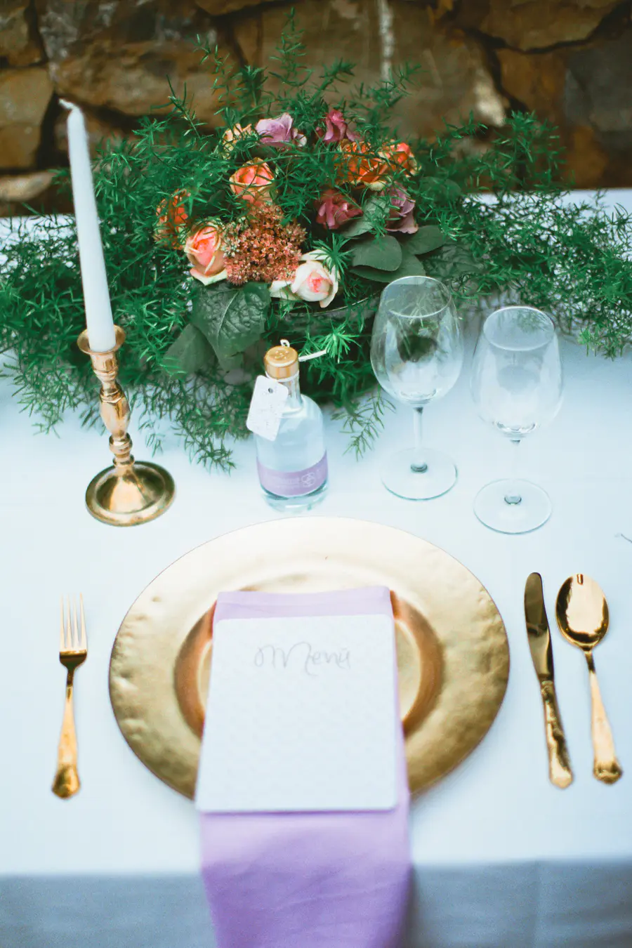 A beautifully arranged table setting featuring a gold charger plate, a lavender napkin with a "Menu" card, and gold cutlery. A small bottle with a tag and two empty wine glasses sit nearby, analog soul of Fuji Film PRO 400H by Marek and Klara