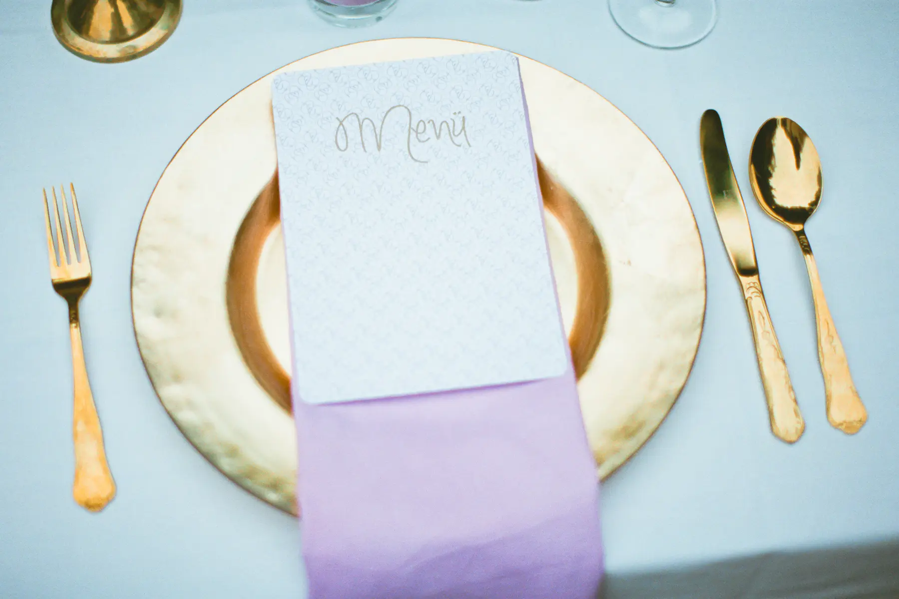 
A formal place setting on a light tablecloth, featuring a gold charger plate, a folded lavender napkin, and a white menu card with "Menu" written in gold script. A gold fork is to the left, and a gold knife and spoon are to the right.