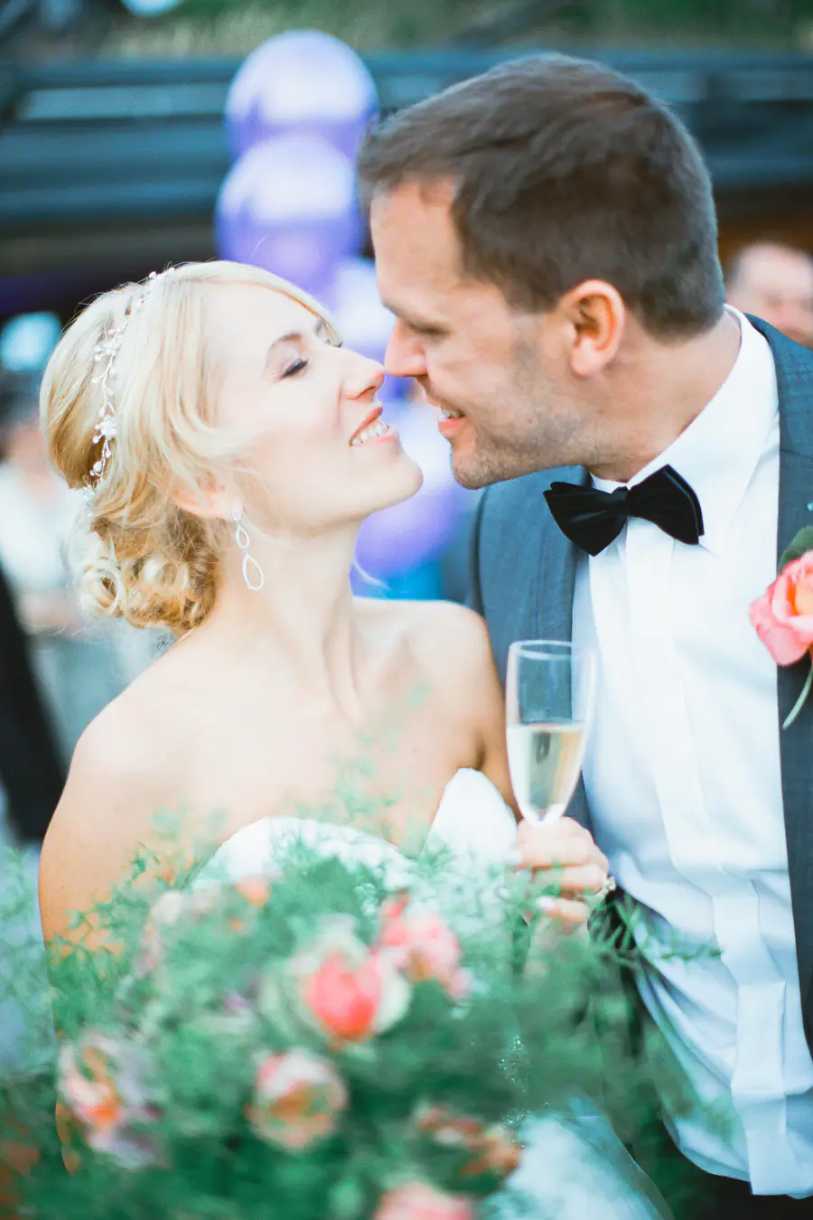 Cantured on Fuji Film PRO 400H by Marek and Klara. A bride and groom in wedding attire affectionately touch noses. The bride wears a strapless dress and a delicate headpiece, holding a bouquet with pink flowers and green foliage. The groom wears
