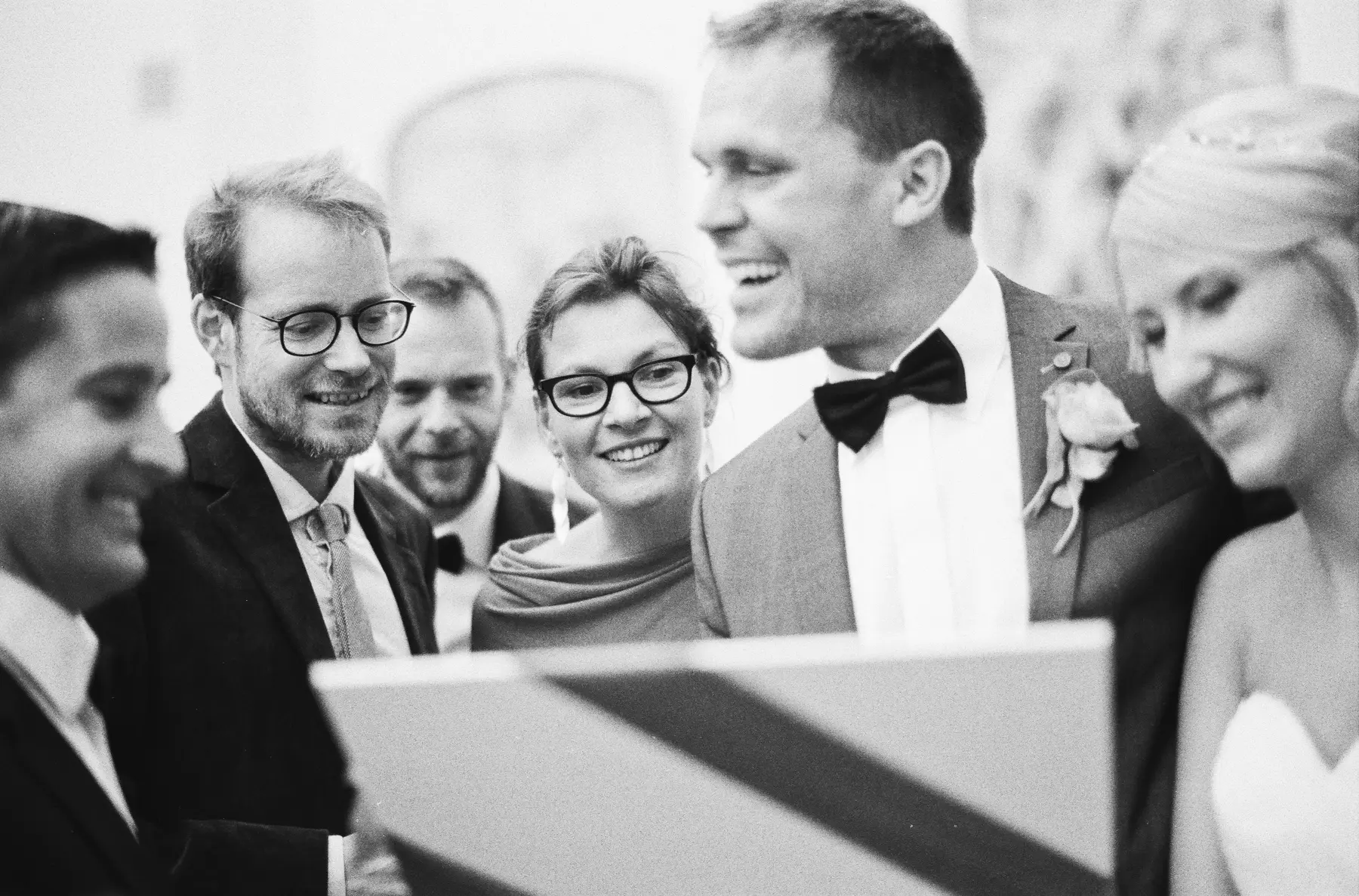 Photojournalistic wedding photography Vienna: A group of people, including a bride and groom, are looking down and smiling at something held in front of them, possibly a tablet or phone, in a black and white photo.