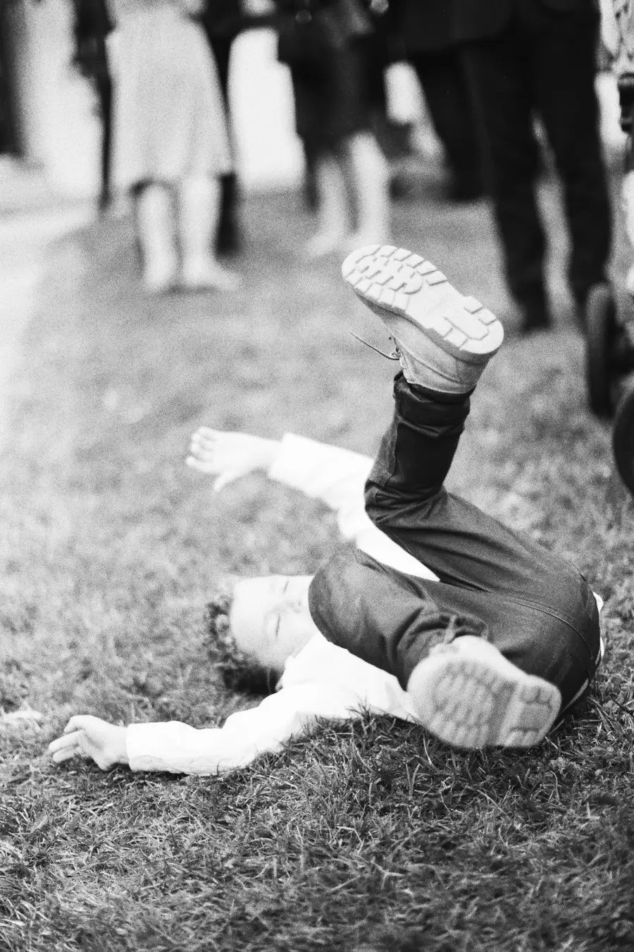 A young child is lying on their back in the grass with their legs in the air, appearing to be playing or falling.