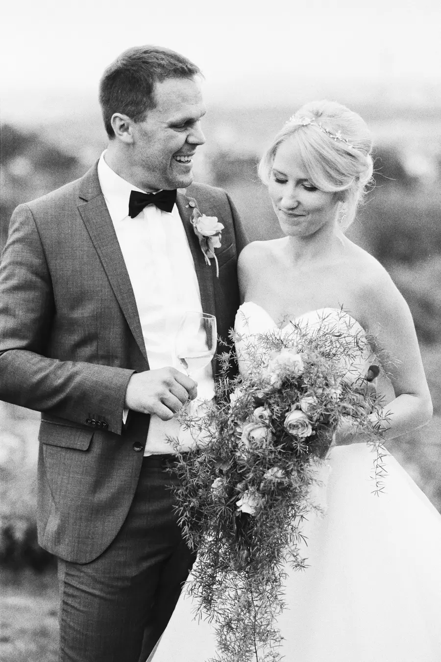 Black and white photo -Kodak Tri-X 400 Pro- of a smiling bride and groom outdoors. The groom, in a suit and bow tie, holds a wine glass and looks happily at the bride, who wears a strapless gown, a ti