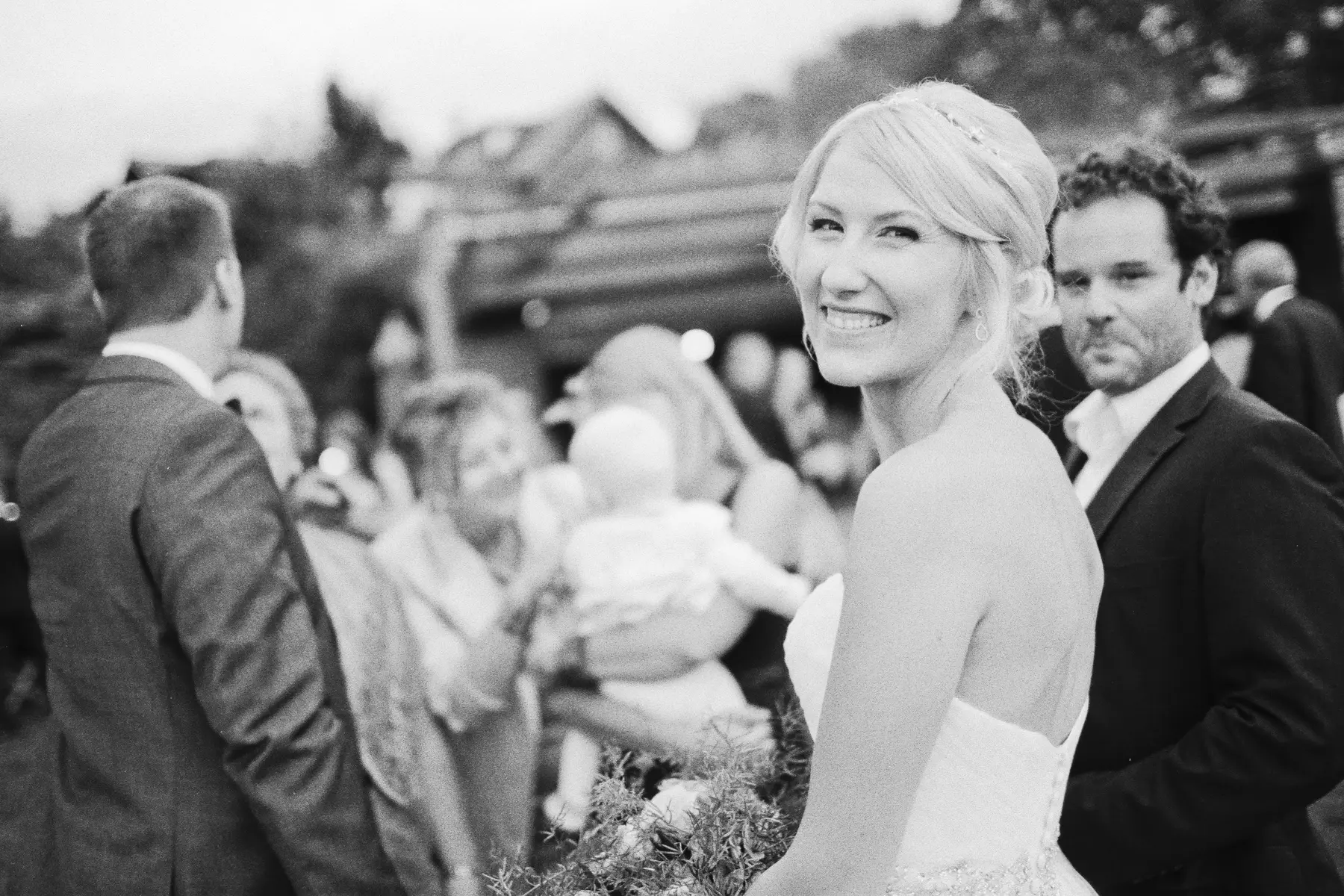 Captured on Kodak Tri-X 400 Pro.A black and white photo of a smiling blonde bride in a strapless wedding dress, looking over her shoulder at the camera, holding a bouquet. Her partner, a man in a dark suit