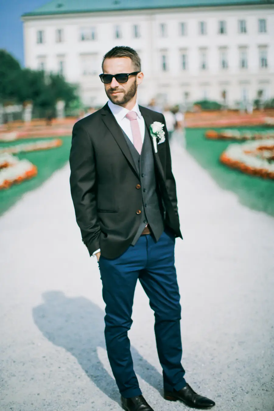 A man in sunglasses, a dark suit jacket, grey vest, white shirt, and pink tie stands with hands in his blue trousers pockets. He has a white boutonnière on his lapel