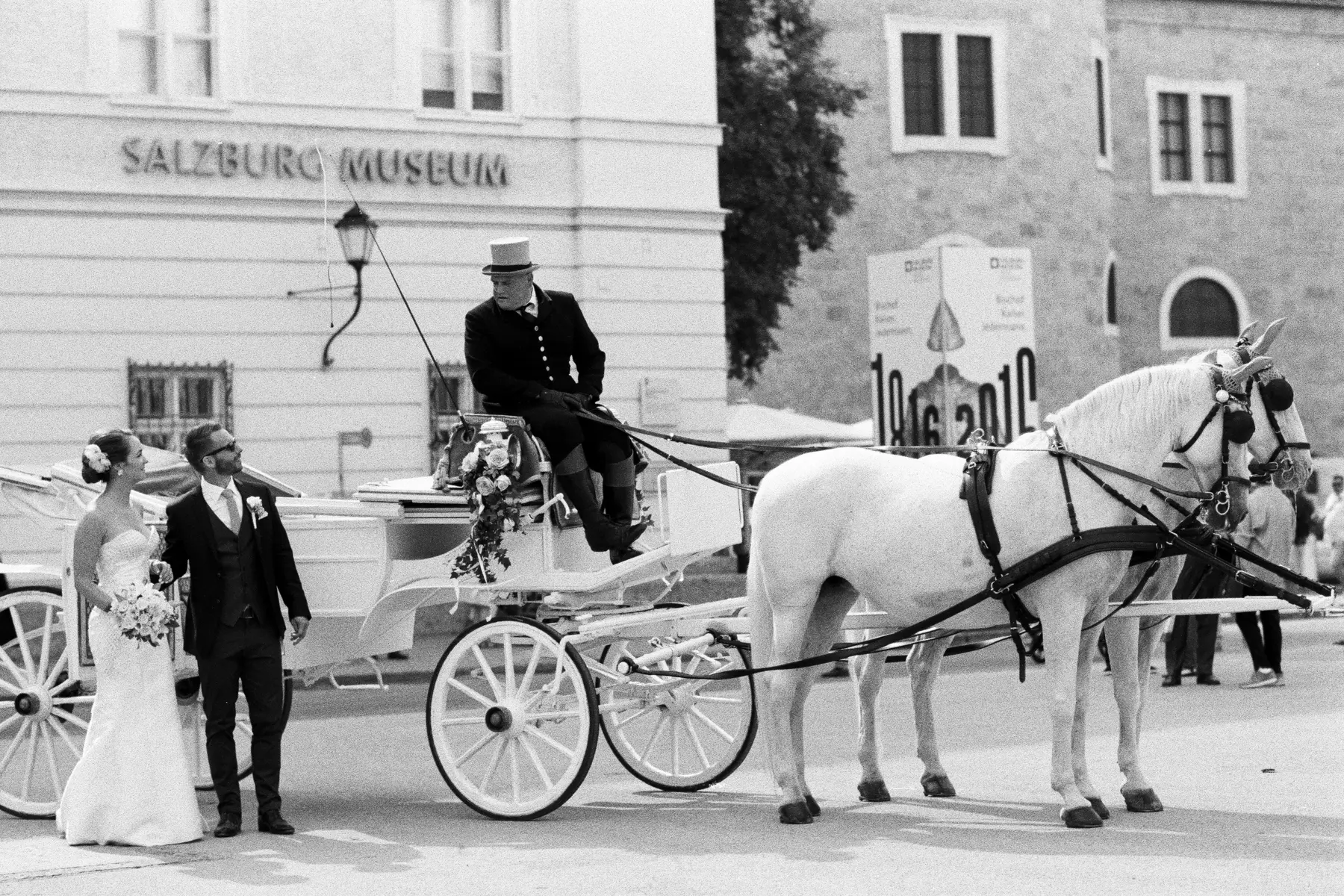 Timeless black and white wedding portrait, shot on Kodak Tri-X 400 film. A bride and groom stand beside a white horse-drawn carriage in front of the Salzburg Museum.