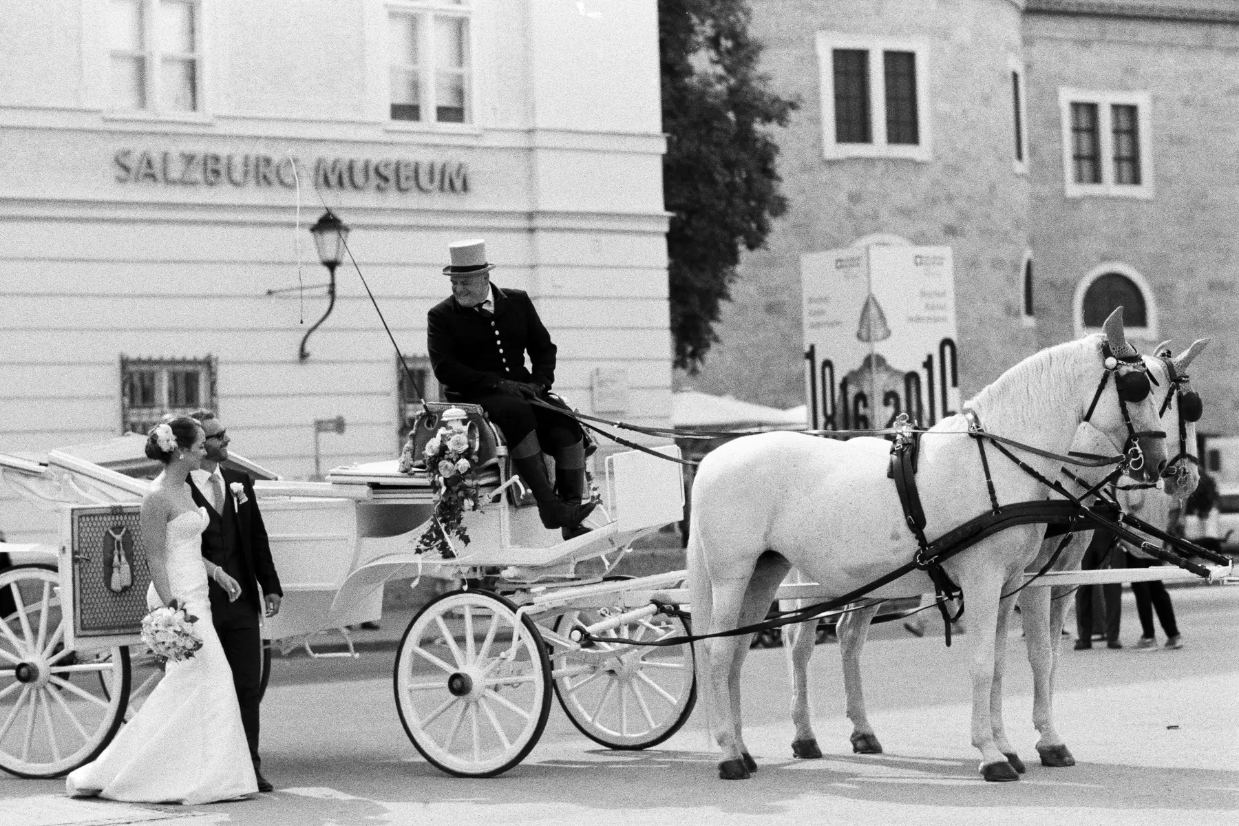 Raw wedding reportage captured on Kodak Tri-X 400 – high-contrast black and white film photography. A bride and groom stand beside a horse-drawn carriage with the Salzburg Museum in the background.