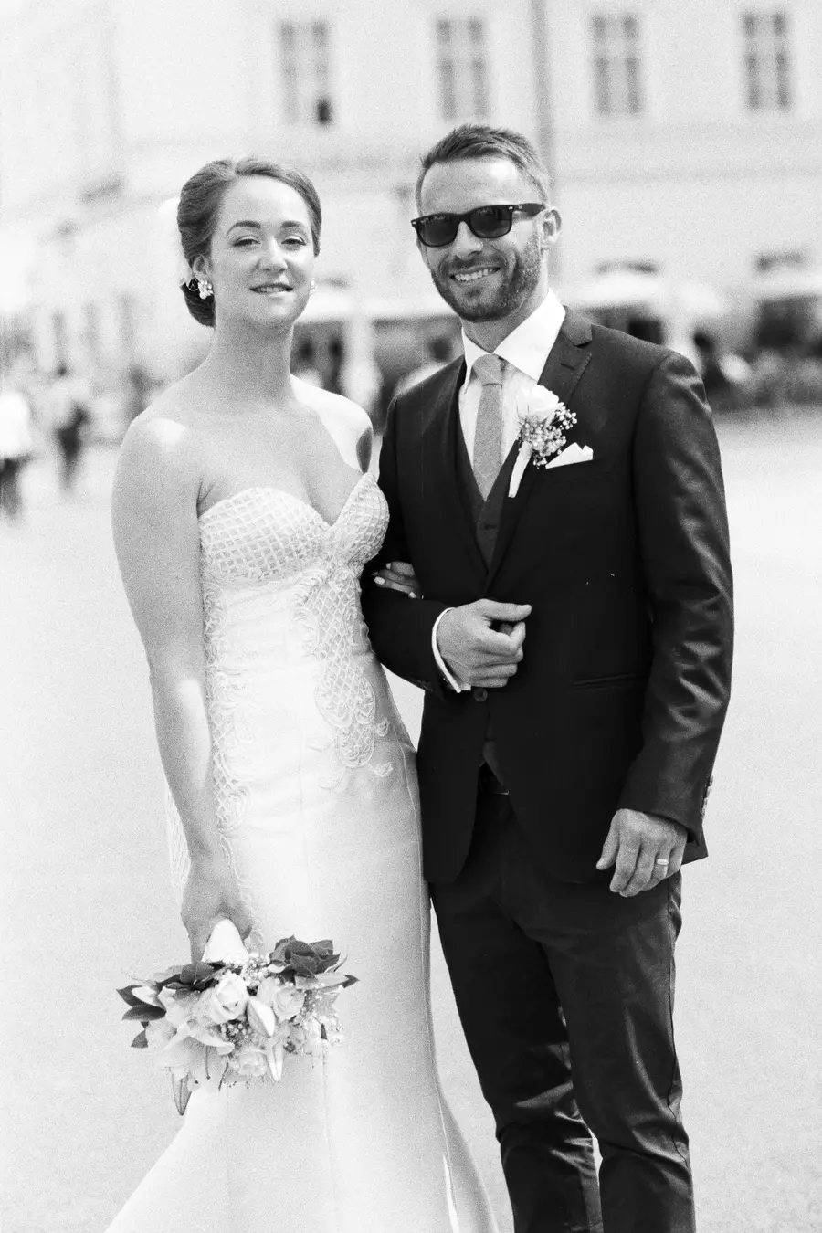 A black and white photo of a smiling bride and groom posing outdoors. The bride, on the left, wears a strapless white wedding gown and holds a bouquet, her arm linked with the groom