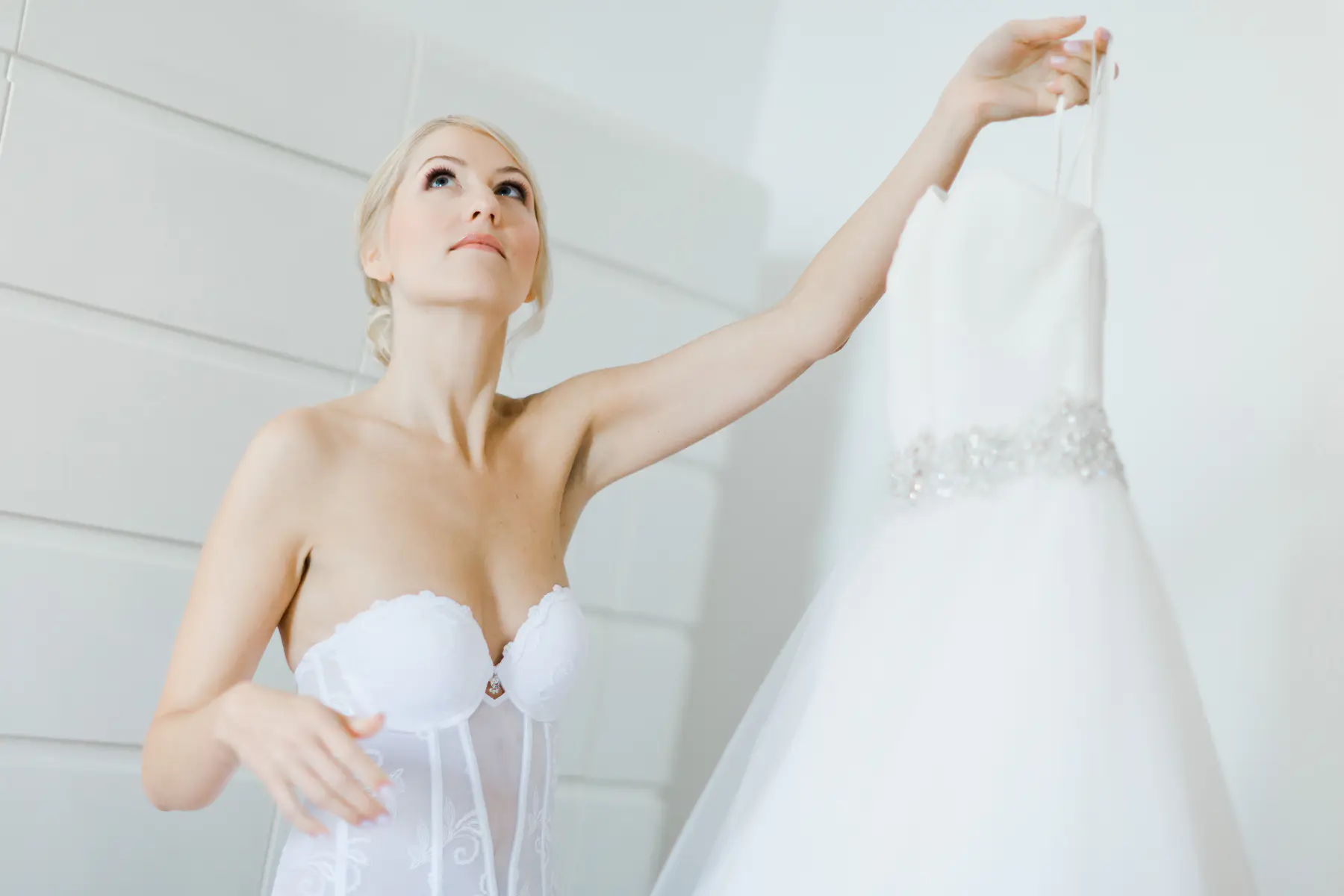 A bride looks up at her white wedding dress as she holds it up by the straps.