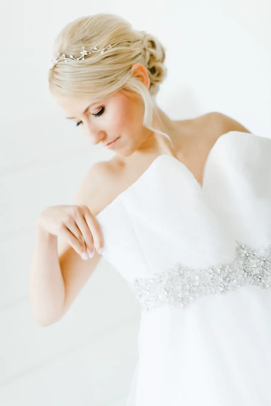 A blonde bride with her hair in a bun and wearing a tiara looks down at her white strapless wedding dress, which has a beaded belt.