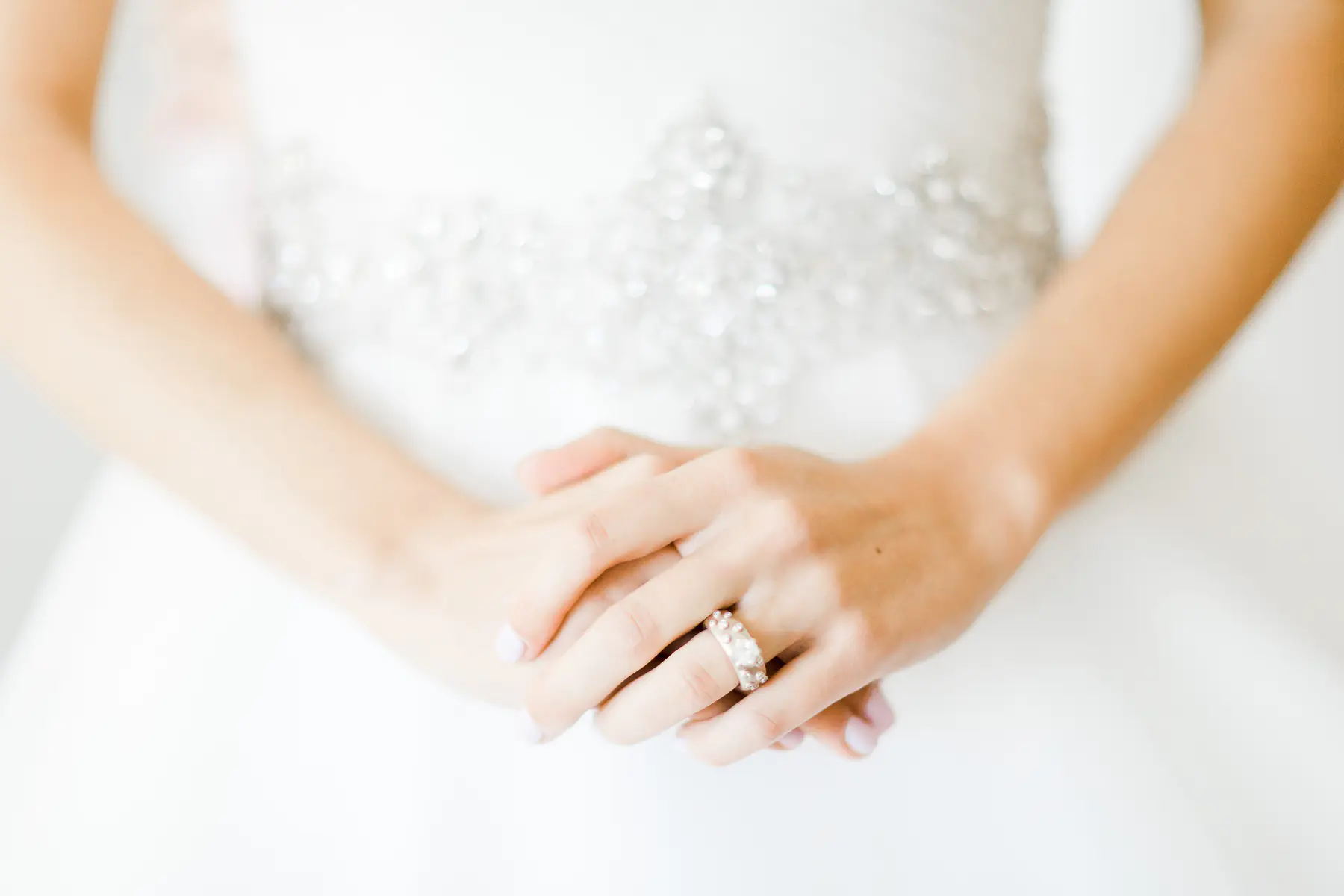 A bride's hands are clasped, with a diamond ring on her left hand. The bride is wearing a white wedding dress with a beaded bodice.