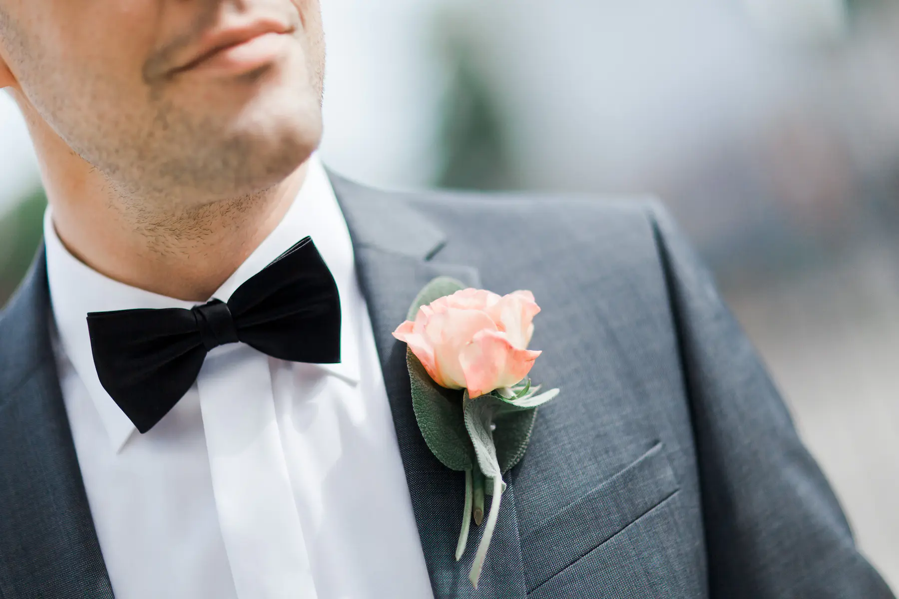 A close-up shot shows the lapel and upper chest of a person wearing a grey suit jacket, a white shirt, and a black bow tie. A boutonnière with a light peach-colored rose and green leaves is pinned to the jacket.
