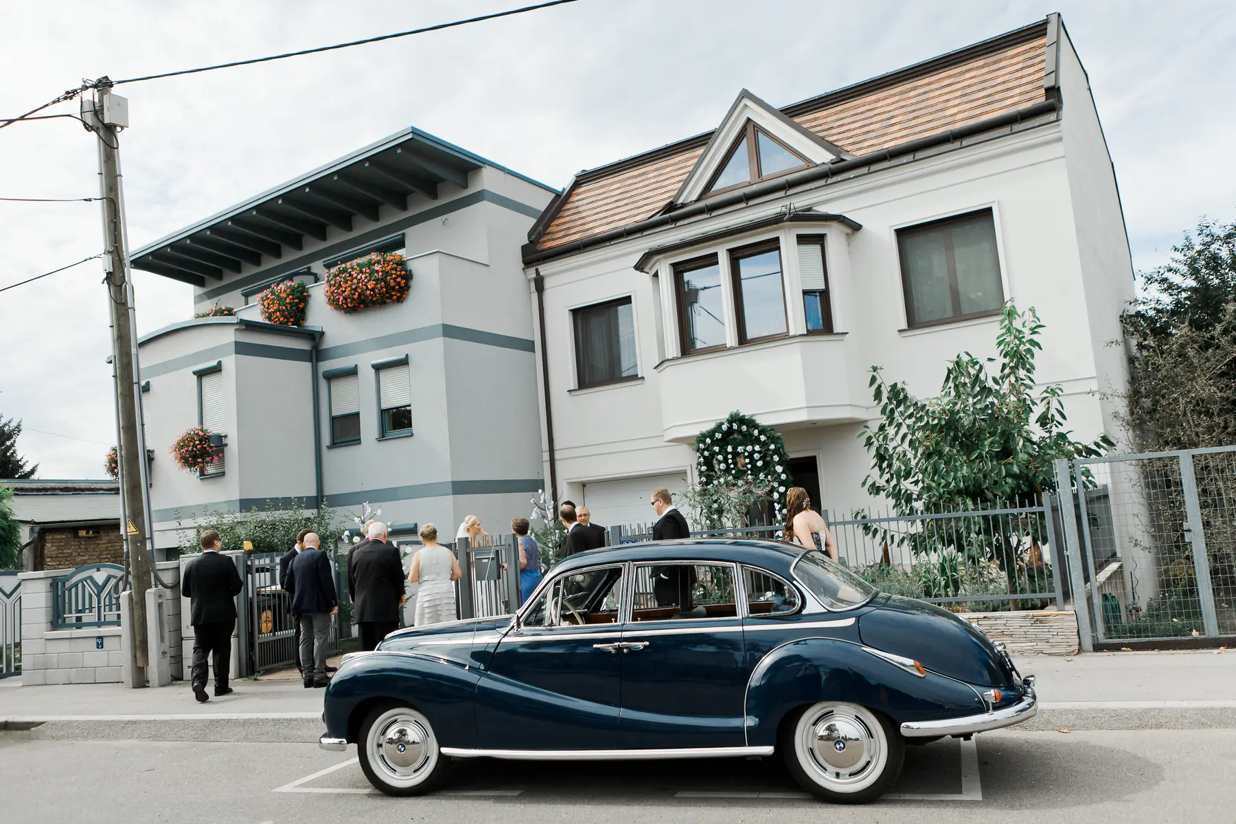 A dark blue vintage car is parked on the street in front of two modern houses. Several people in formal attire are gathered on the sidewalk in front of the houses, suggesting a special occasion.