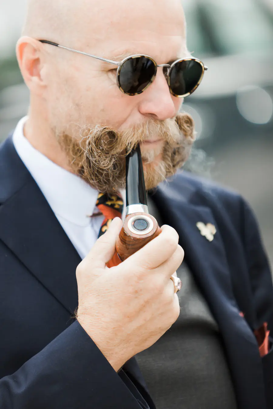 A close-up shot shows a bald man with a light brown mustache and beard, wearing sunglasses and a dark suit. He is smoking a wooden pipe.