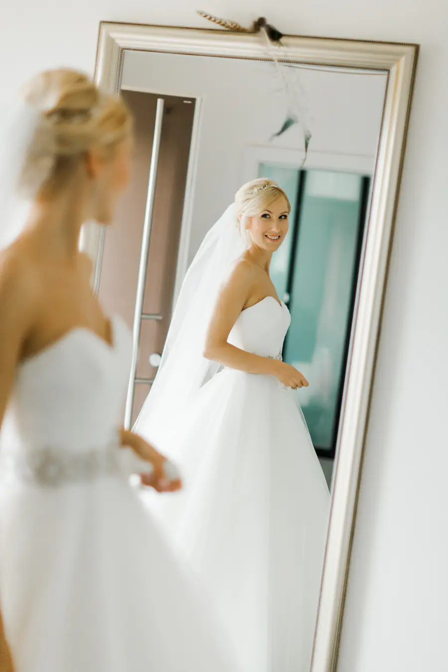 A bride in a white wedding dress smiles at her reflection in a large mirror.
