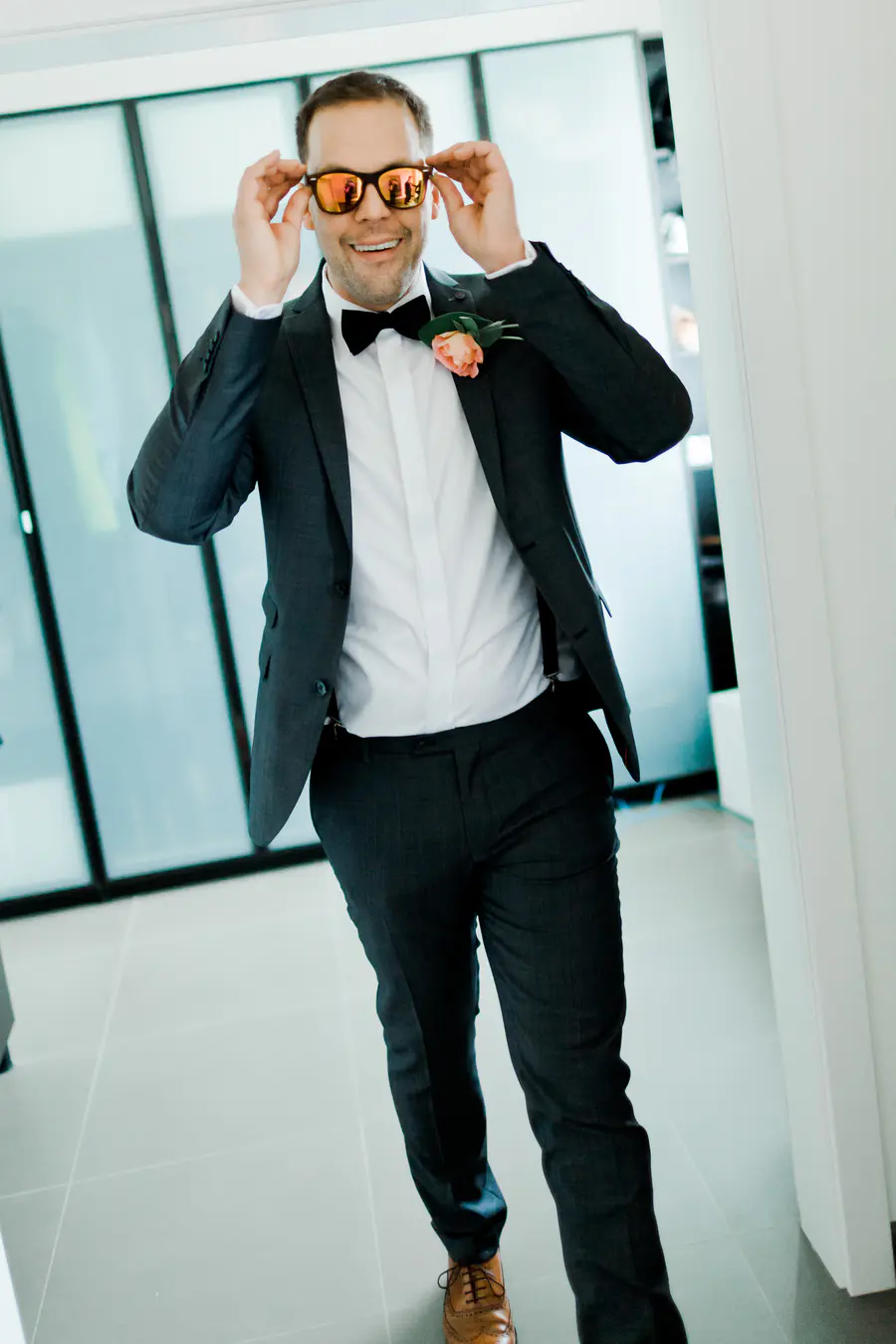 Candid wedding photo: A man in a dark suit, white shirt, bow tie, and suspenders adjusts orange-tinted sunglasses. A peach rose boutonniere is pinned to his lapel.