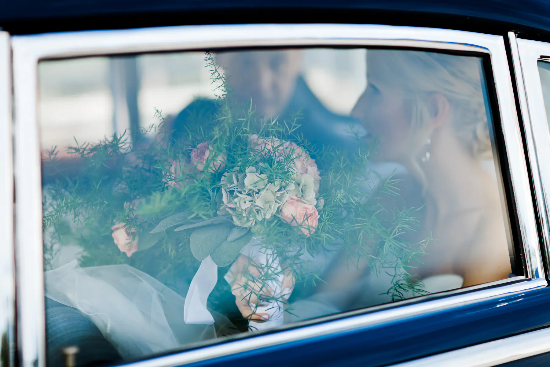 A bride and groom are reflected in the window of a car, with a wedding bouquet in the foreground.