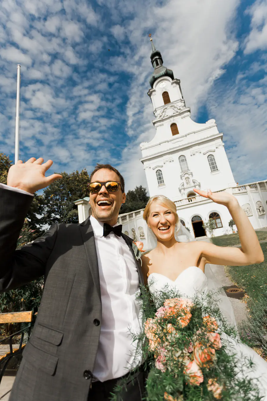 Marek & Klara Wedding photo Vienna: A happy bride and groom wave at the camera in front of a white church with a steeple. The groom wears a suit and sunglasses, while the bride is in her wedding dress holding a bouquet.