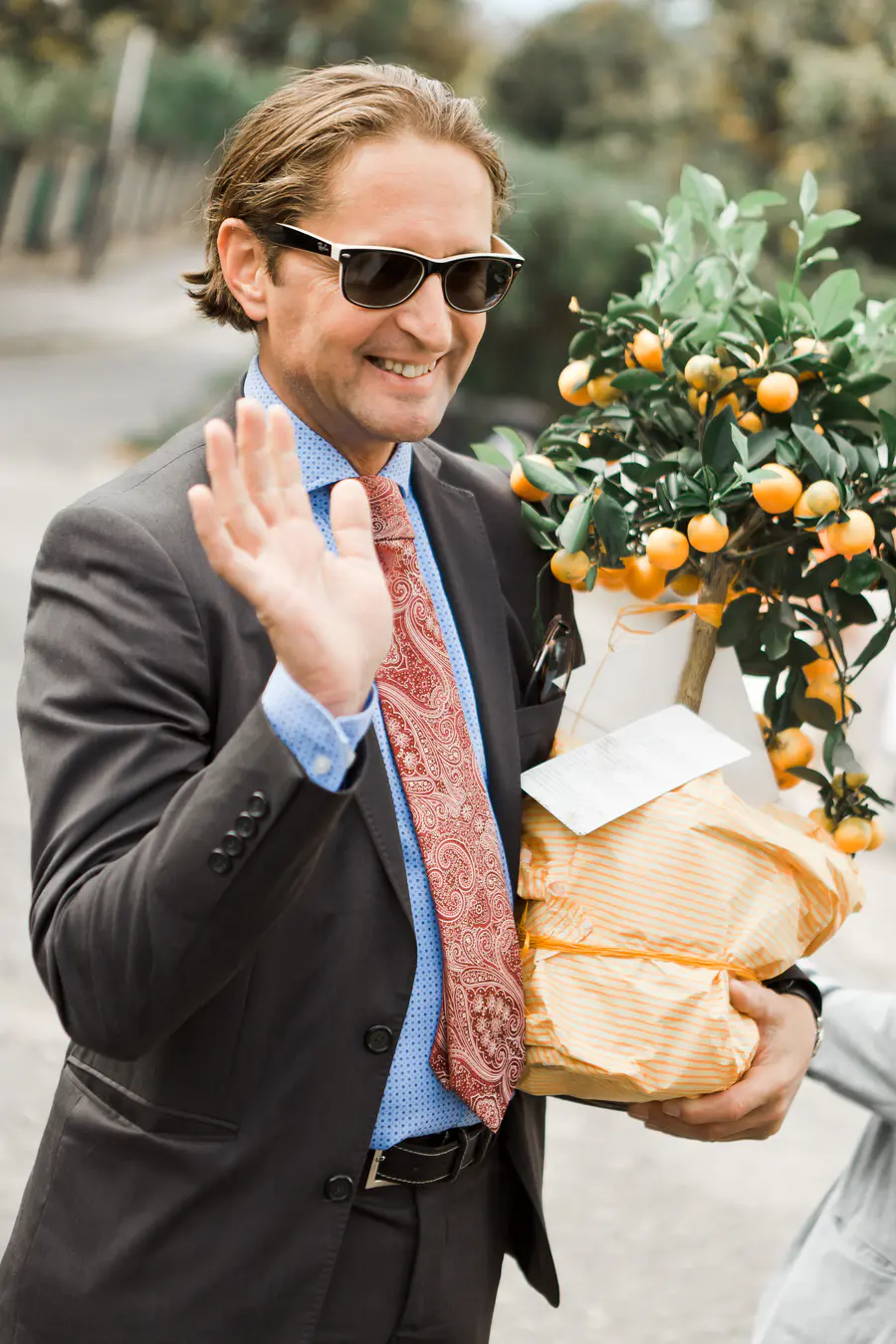 A man in a suit and sunglasses waves while holding a small potted tree with orange fruits and a wrapped gift.