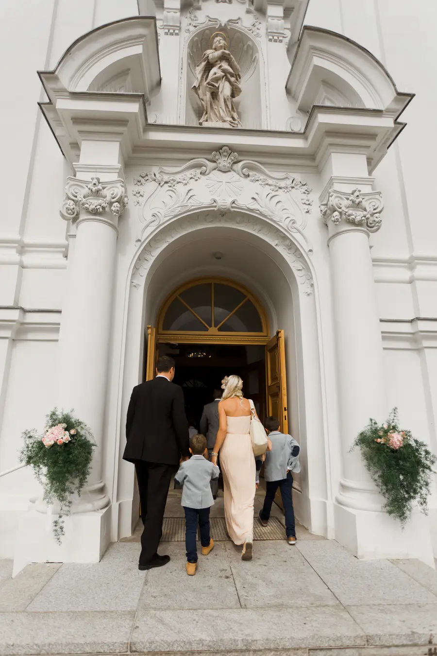 A wedding party walks into a white church through a large arched doorway.