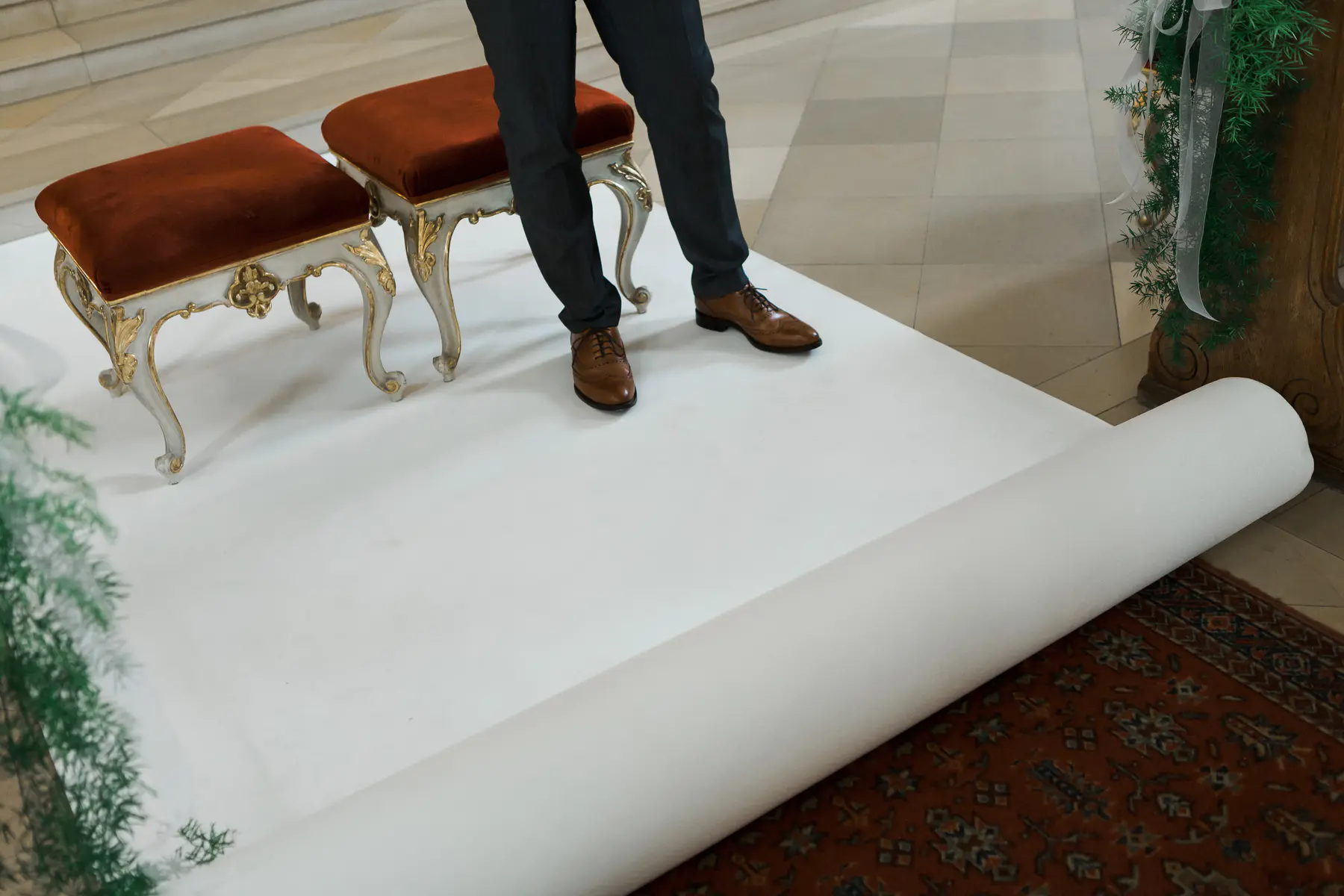 A person in a suit stands on a white carpet runner that is unrolled, with two ornate red velvet stools beside them.