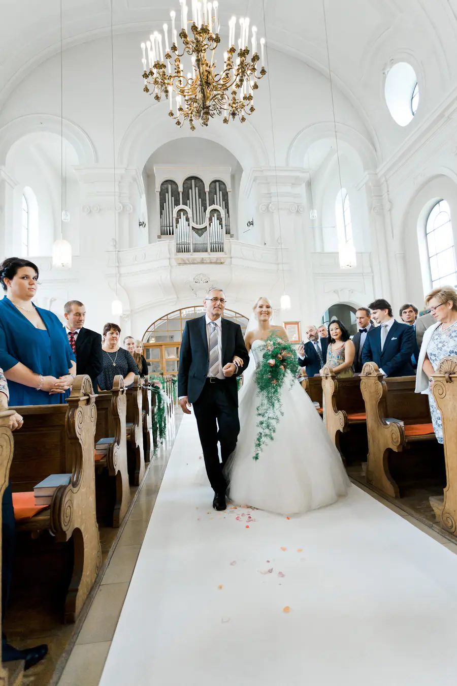 A bride in a white gown is walked down an aisle by an older man in a suit, while guests watch from pews in a church.