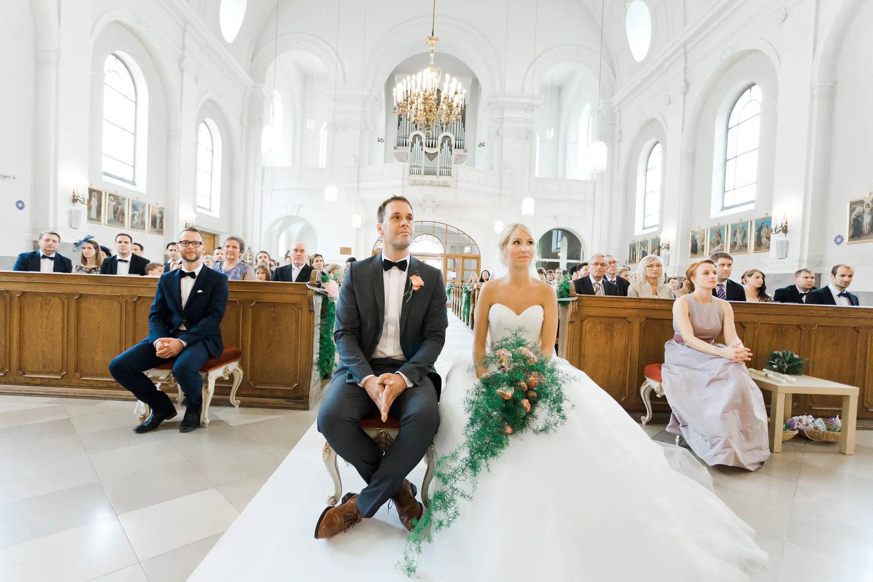 A bride and groom sit in the front pews of a church, looking forward. The groom is in a dark suit with a bowtie and a boutonnière, while the bride wears a white strapless wedding gown. Guests are seated in the pews behind them, and an organ is visible in the background.