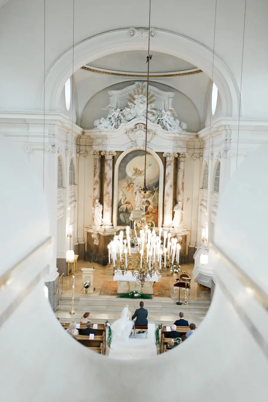 A bride and groom sit at the altar in a bright, ornate church during a wedding ceremony.