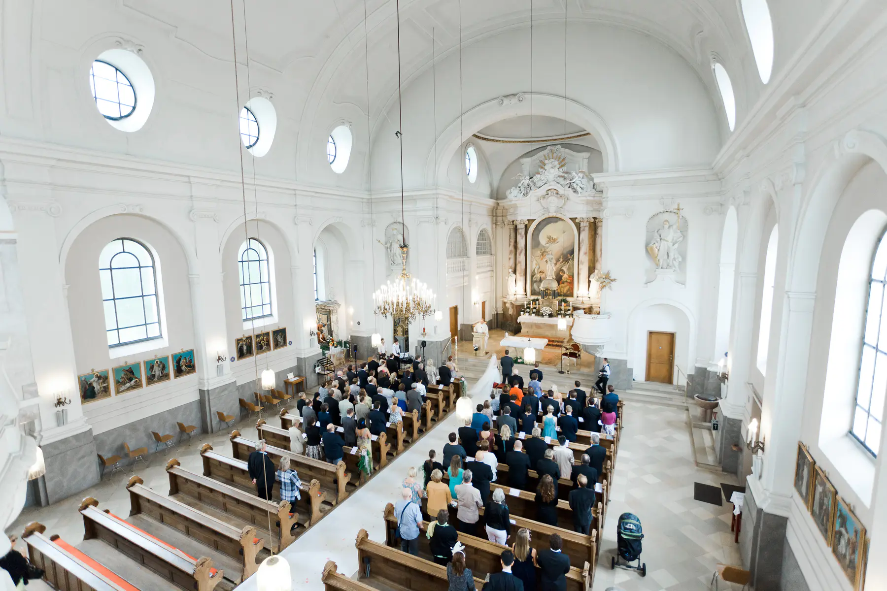 A high-angle shot shows a wedding ceremony taking place in a brightly lit, white church. Guests are seated on wooden pews, and a wedding party is gathered at the altar.