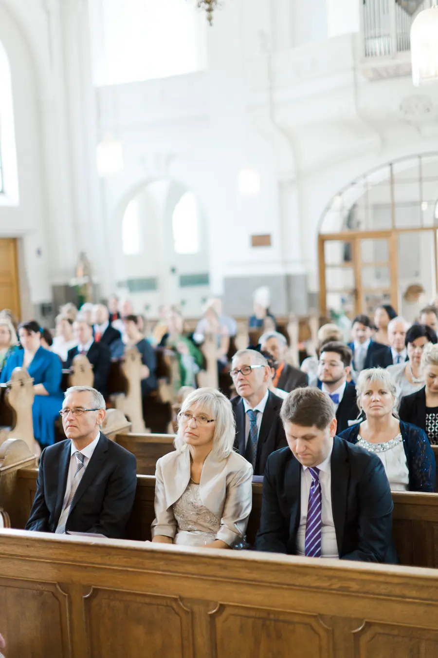 A diverse group of people, dressed in formal attire, sit in pews inside a bright, airy church.