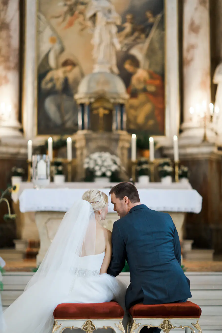 A bride and groom, seen from behind, sit on velvet stools in front of an altar in a church.