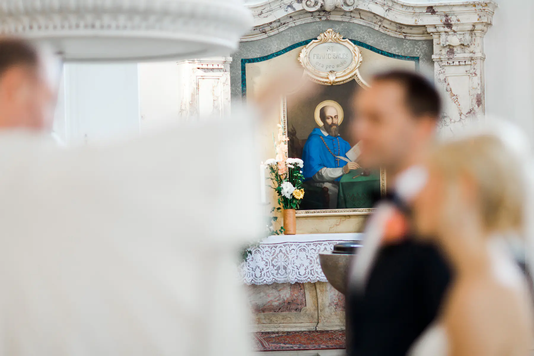 A painting of Saint Francis de Sales is behind a blurred couple in a church.