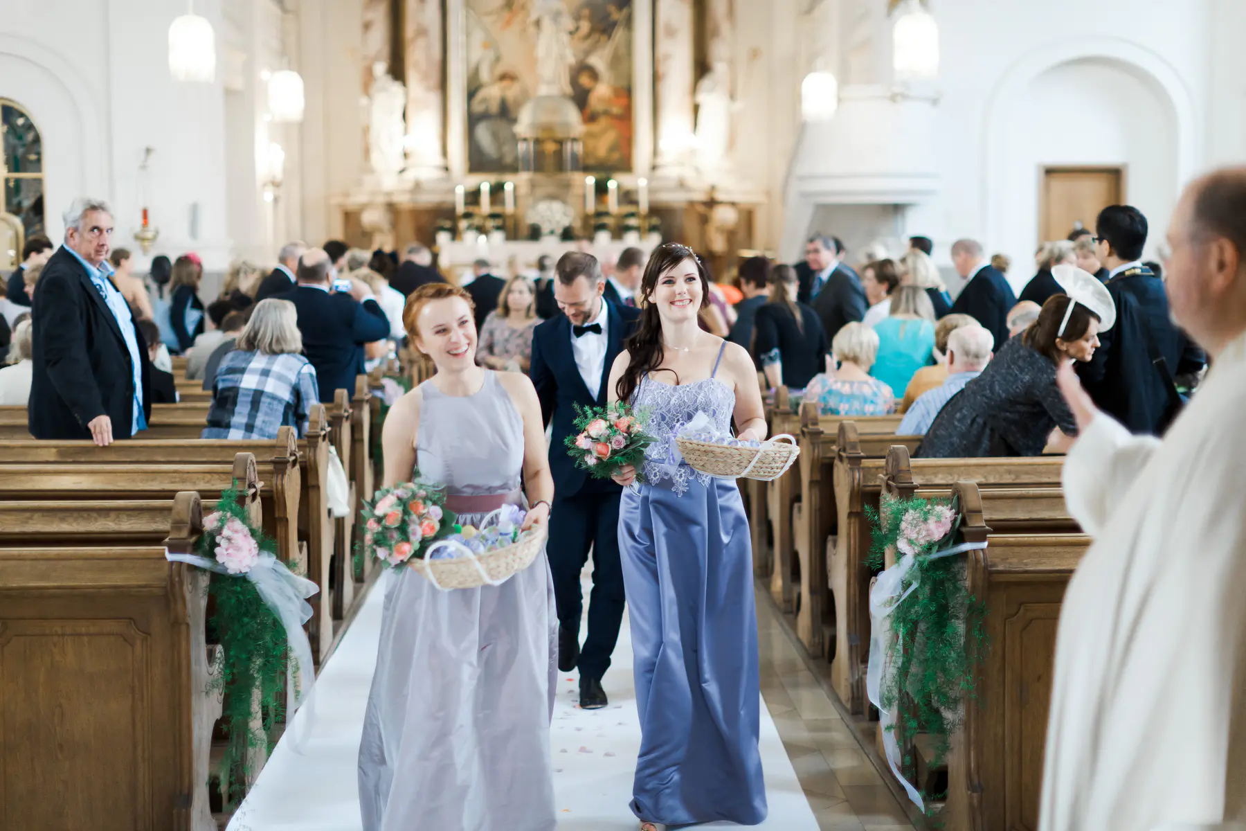 A wedding party walks down the aisle of a church. Two women in lavender dresses are in the foreground, one holding a bouquet and the other a basket. Behind them, a man in a tuxedo follows, and guests are seated in the pews on either side of the aisle.