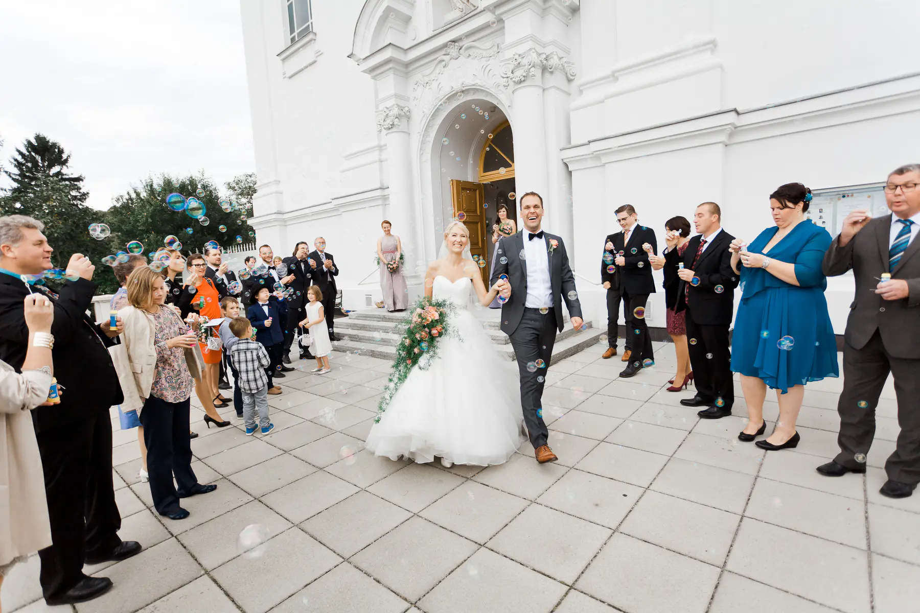 A wedding couple smiles as they walk out of a white building, with guests blowing bubbles around them.