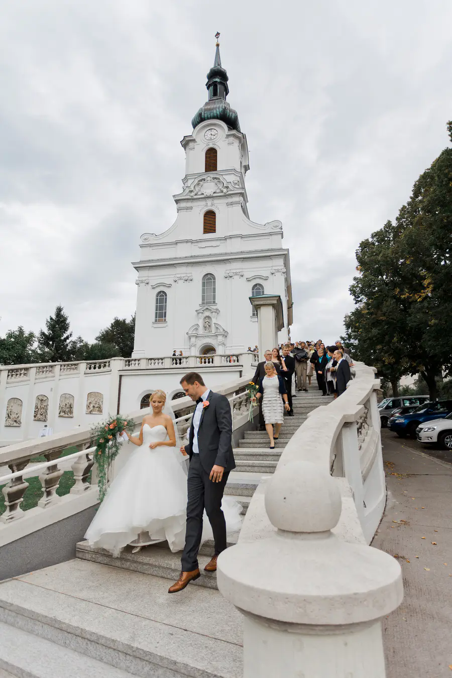 A bride and groom descend a grand staircase in front of a white Kaasgrabenkirche church. The bride wears a strapless white gown and carries a bouquet. The groom is in a dark suit with a bowtie. A wedding party follows them up the stairs.