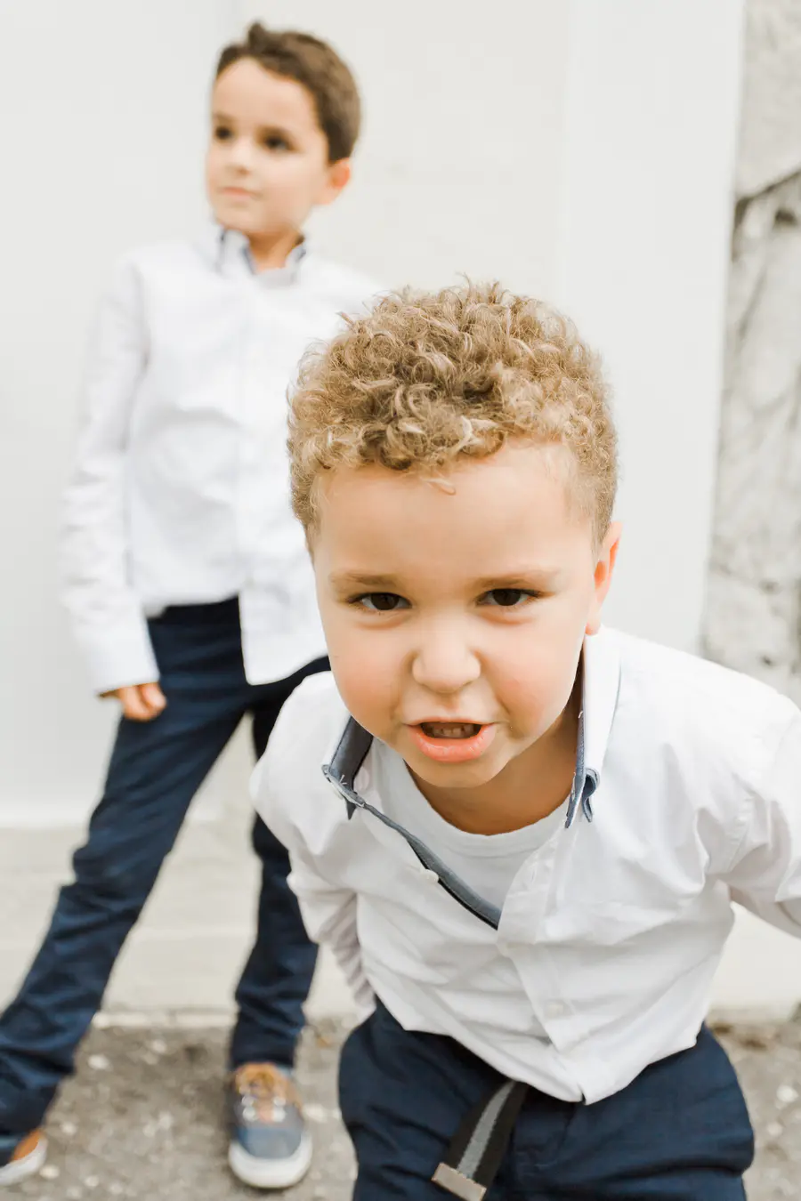 Two young boys in white collared shirts and dark pants. The boy in the foreground has curly blonde hair and is looking directly at the camera with a grimace. The boy in the background is out of focus, facing away from the camera.