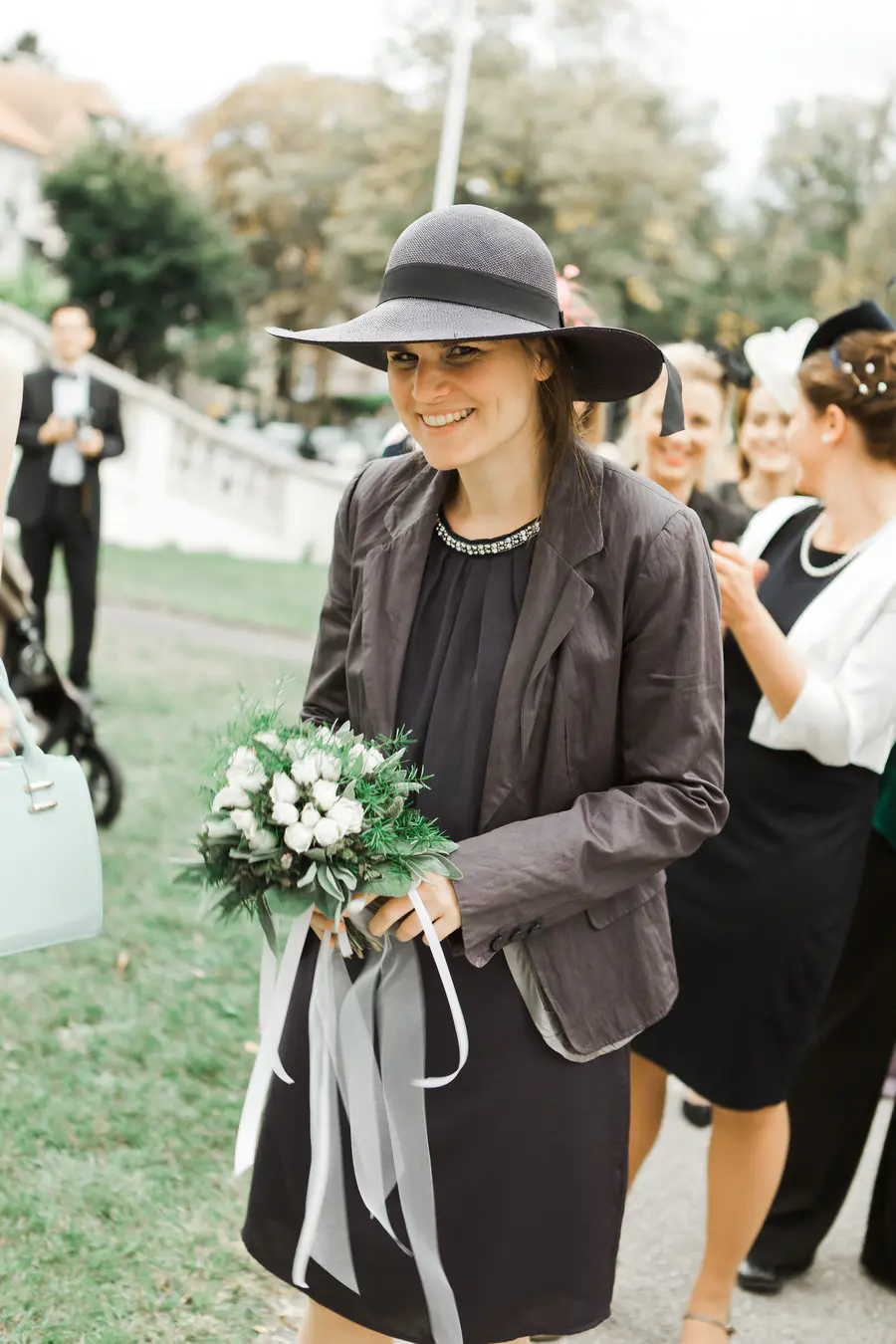 A woman wearing a dark hat and jacket smiles at the camera, holding a bouquet of white roses with flowing white ribbons.