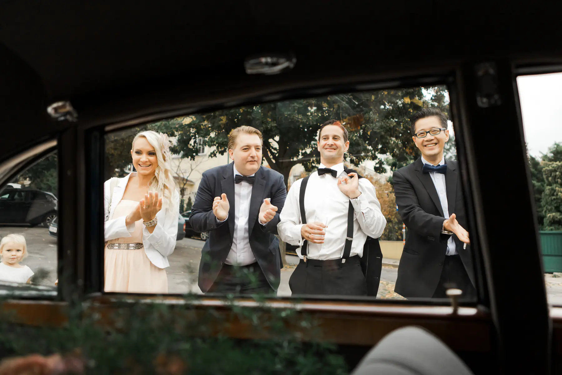 A wedding party of four, two men in tuxedos and two women in formal attire, clap for a young girl dressed in white who is partially visible inside a vintage car.
