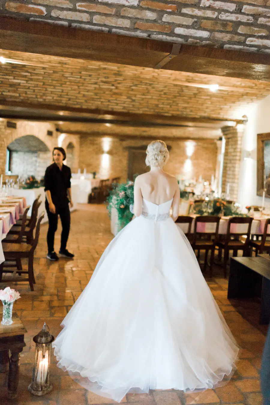 A bride, seen from behind in a strapless white ball gown with a sparkly silver belt, stands in a rustic event hall with brick walls and tiled floors. Tables are set for a reception,