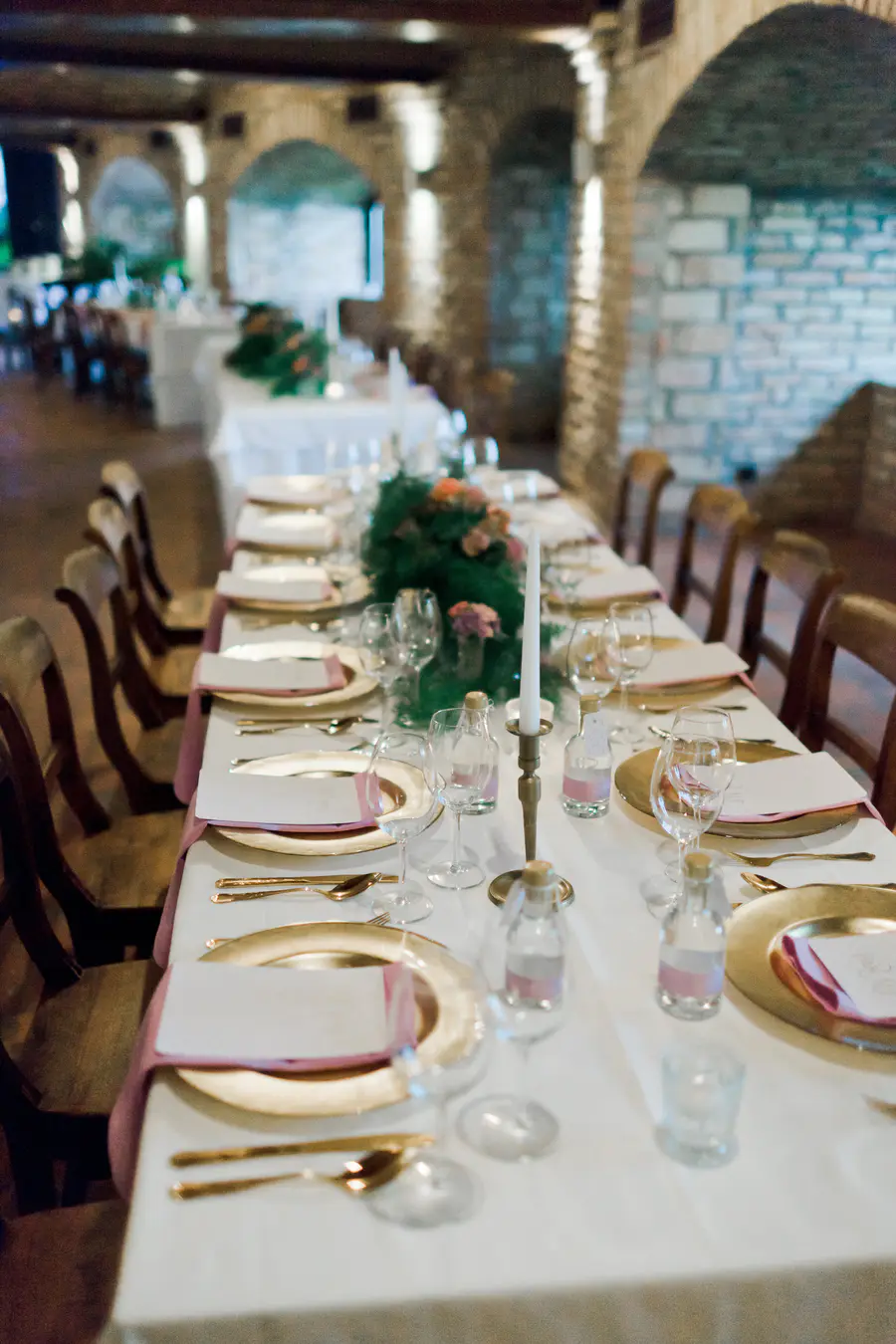 An elegant dining table, set with gold charger plates, white napkins on a pink runner, gold cutlery, wine glasses, and centerpieces featuring green foliage, pink flowers, and tall white candles. The