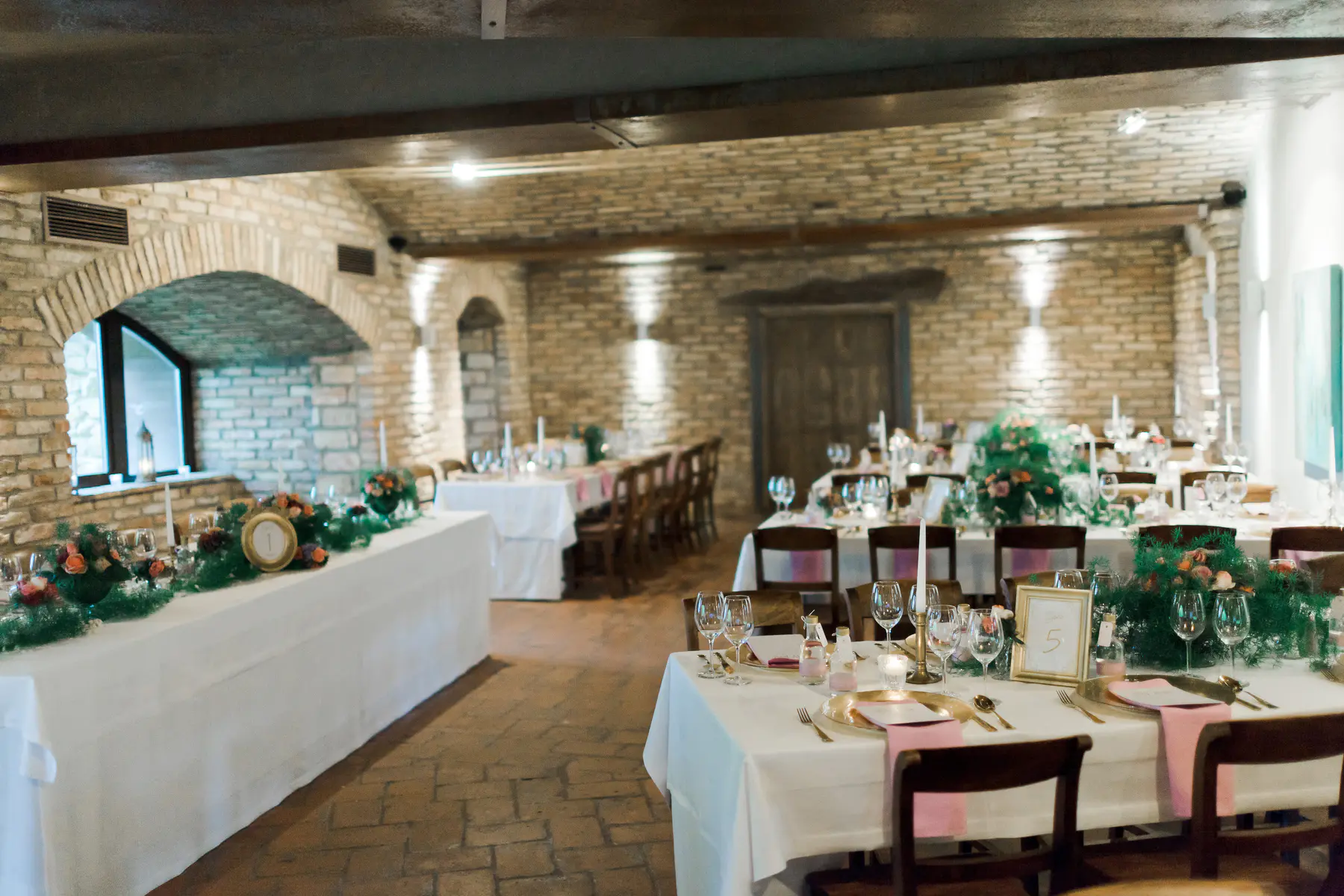 A banquet hall with rustic brick walls and arched windows is set up for a wedding reception. Several long tables are draped in white tablecloths and decorated with greenery, flowers, and candles. Each place setting includes gold chargers, glassware, and pink napkins. A framed table number "5" is visible on one of the tables.