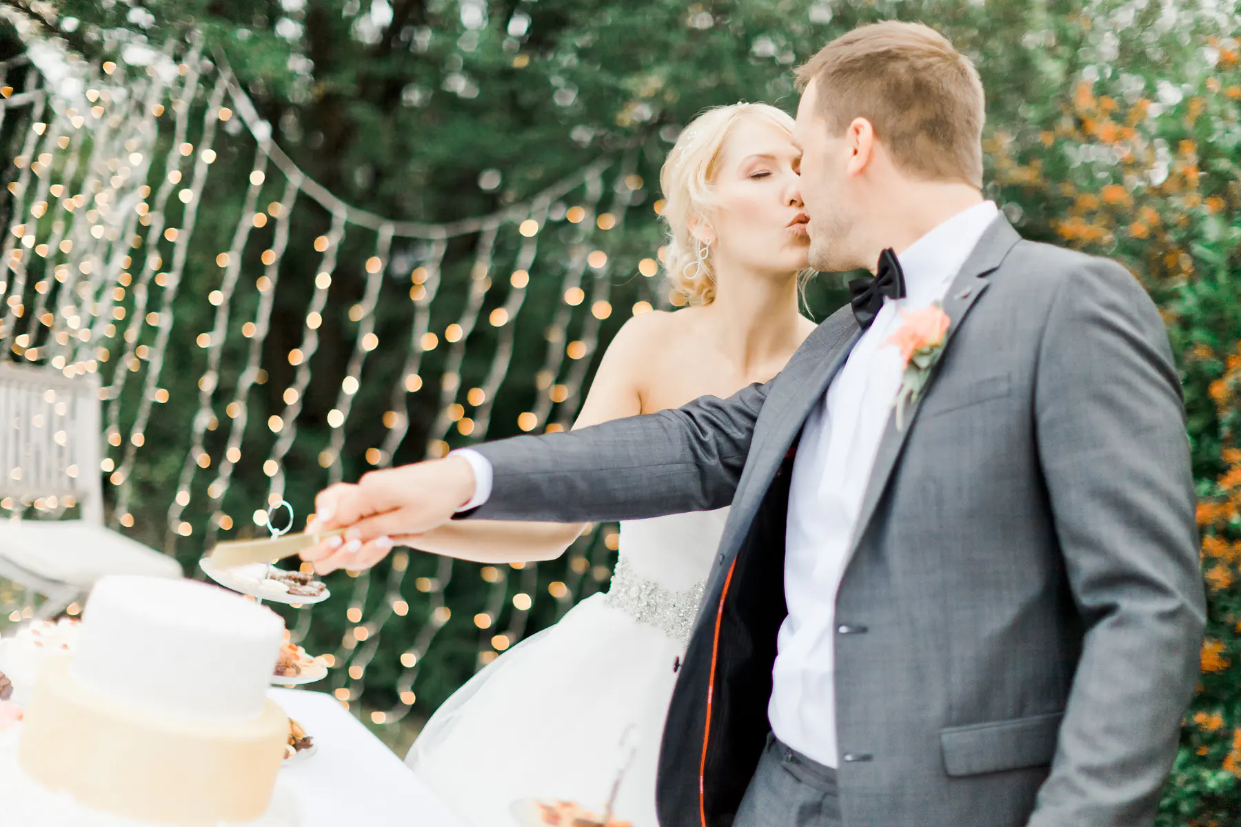 A blonde bride in a strapless white gown and a groom in a grey suit kiss while serving a piece of cake at their outdoor wedding reception. String lights are visible in the blurred green background.